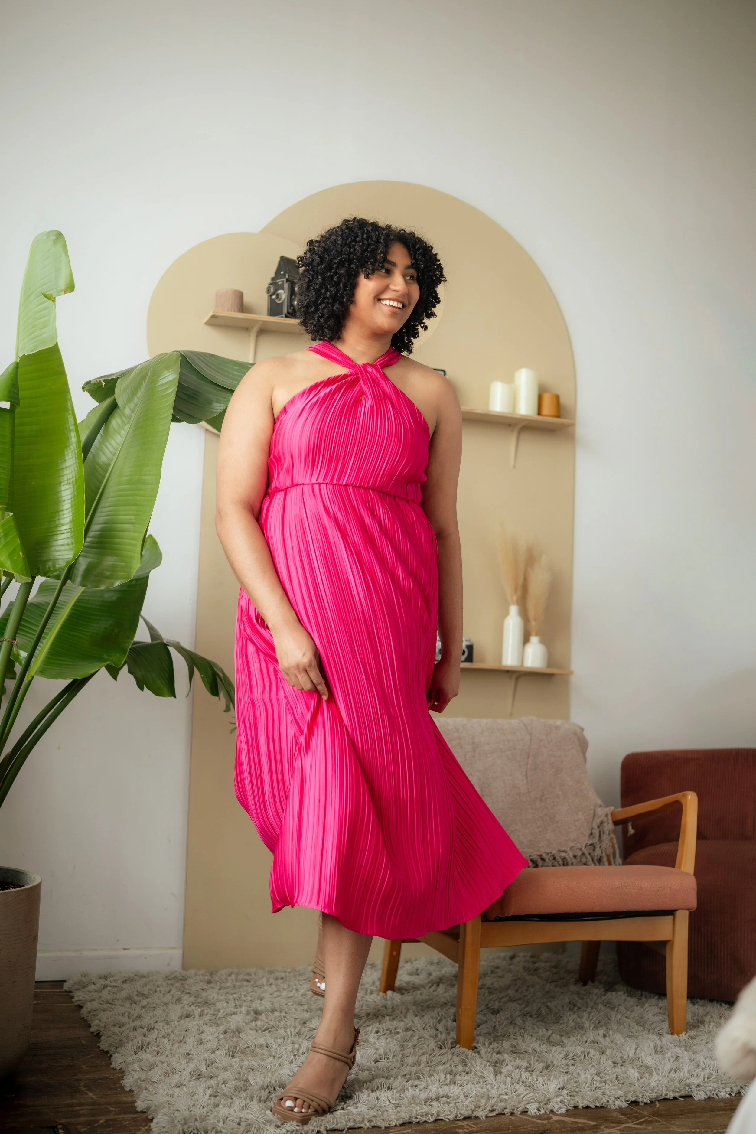 A woman in a bright pink pleated dress standing in a living room, smiling, with a large green plant, beige wall decor, and a beige armchair in the background.