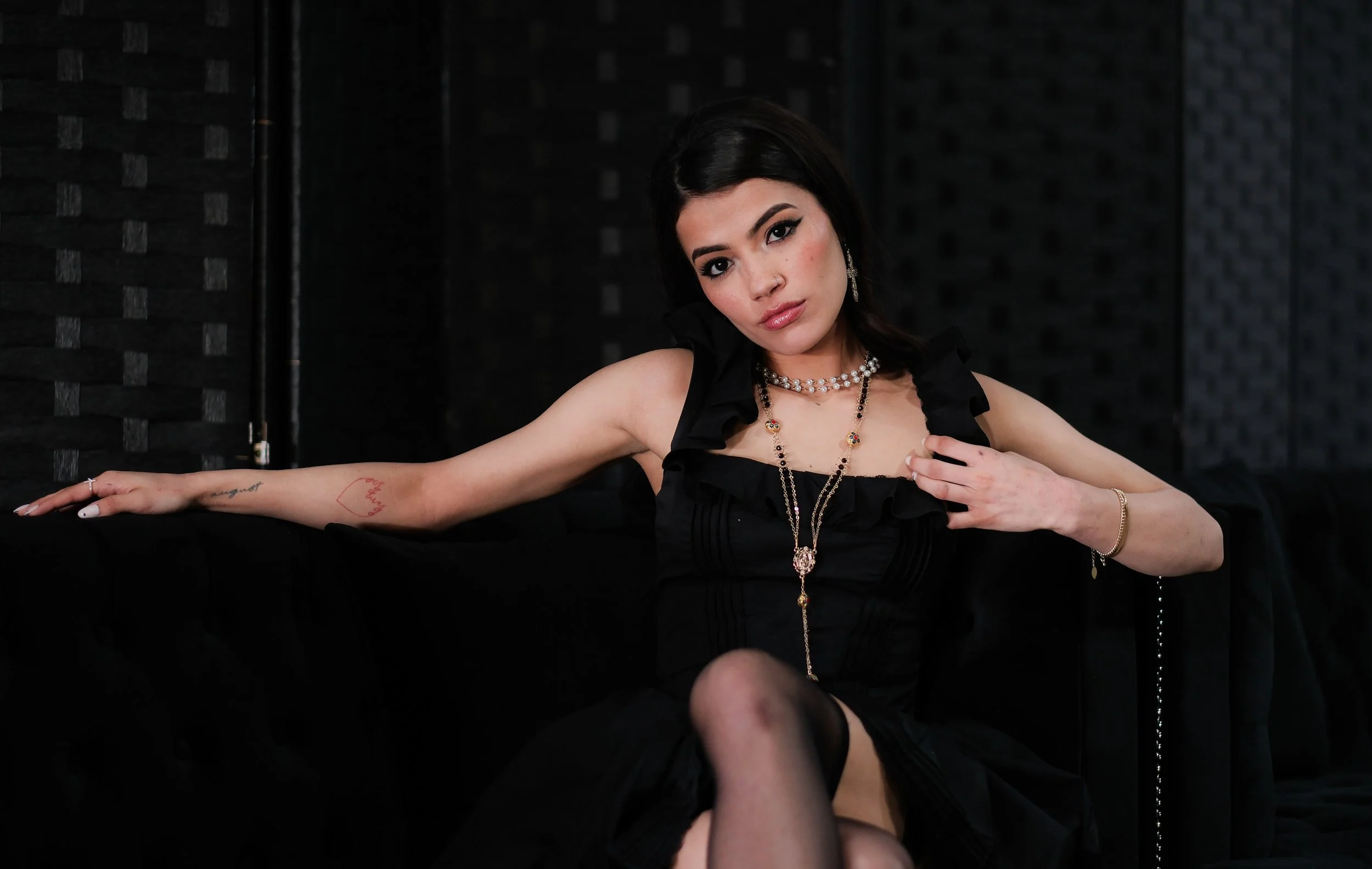 A woman in a black dress sitting on a dark couch with dark walls behind her, looking at the camera.