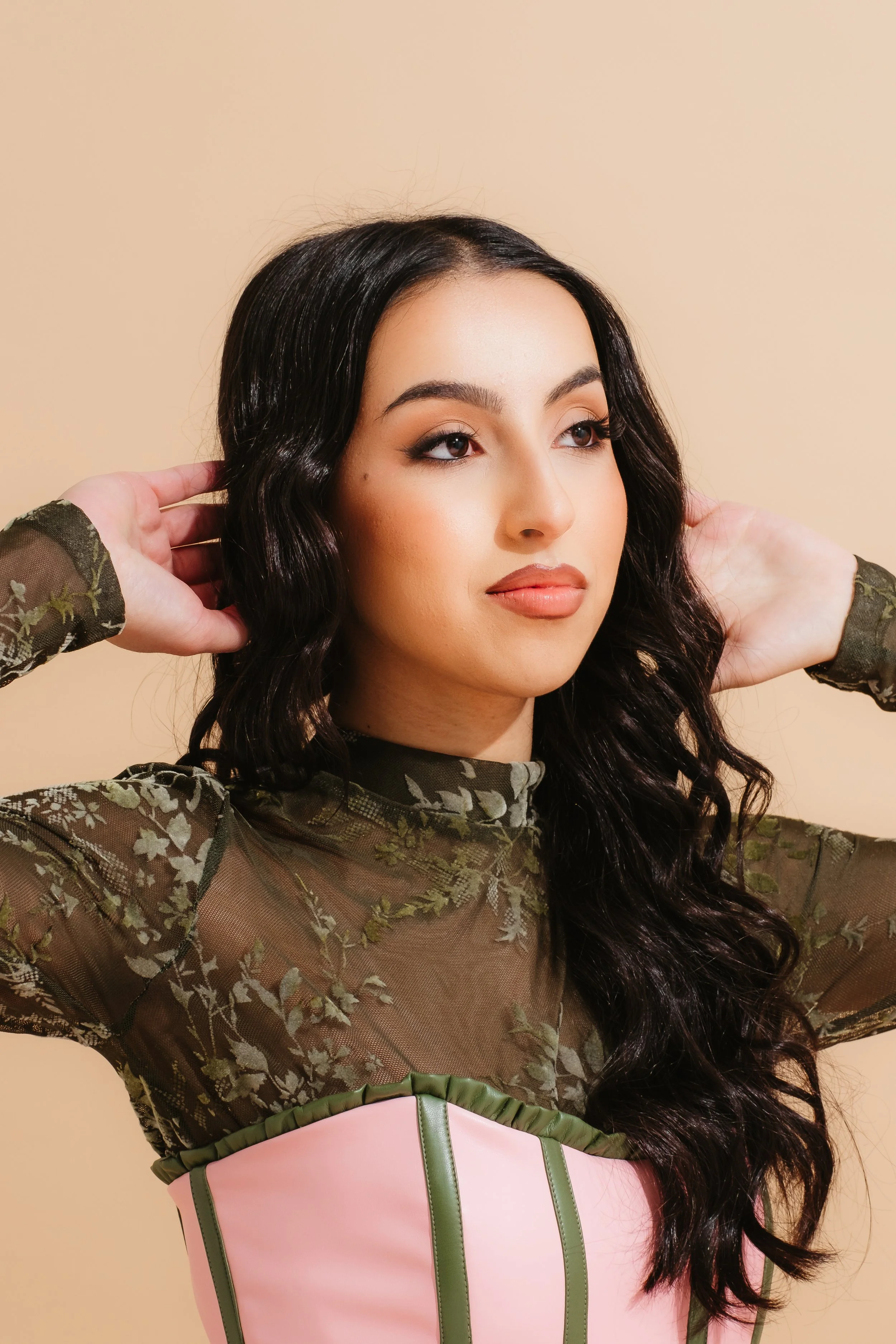 A woman with long dark hair styled in loose waves, wearing a sheer green floral top with long sleeves and a light pink corset with green stripes, standing against a beige background.