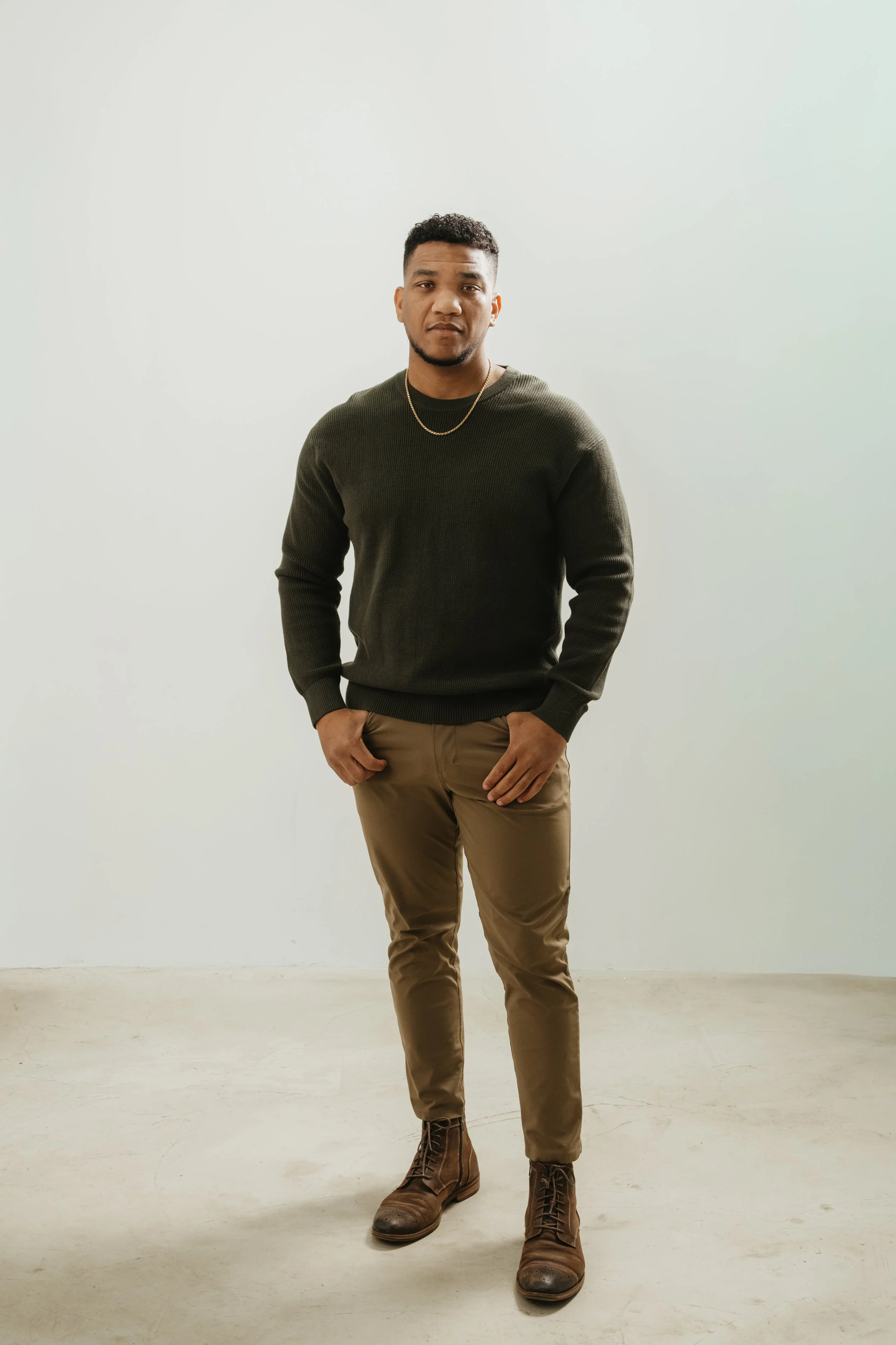 A young man standing against a light-colored wall, wearing a dark sweater, khaki pants, and brown boots, with a gold chain around his neck.