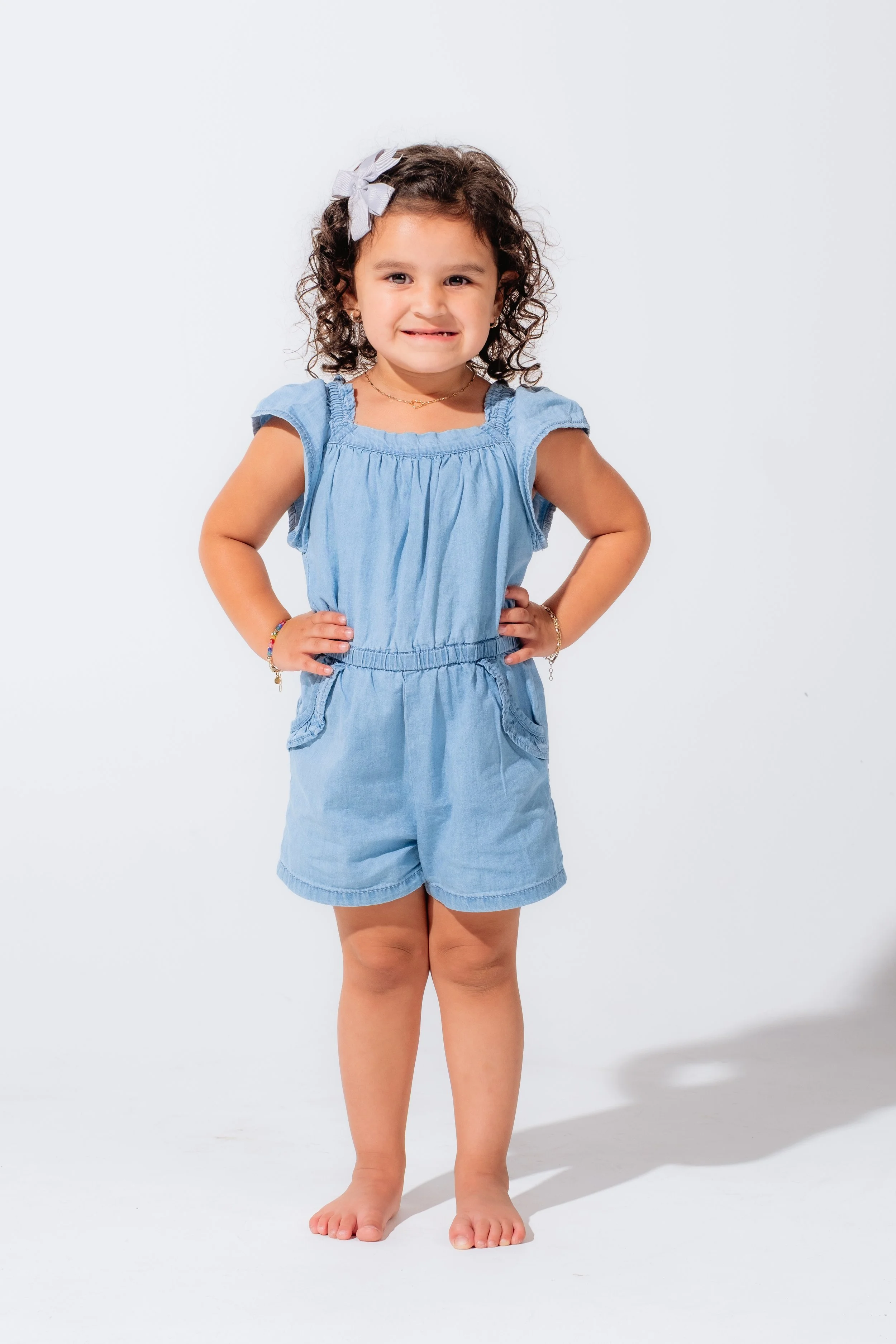 A young girl with curly brown hair, wearing a light blue romper with ruffled sleeves, standing with hands on her hips against a white background.