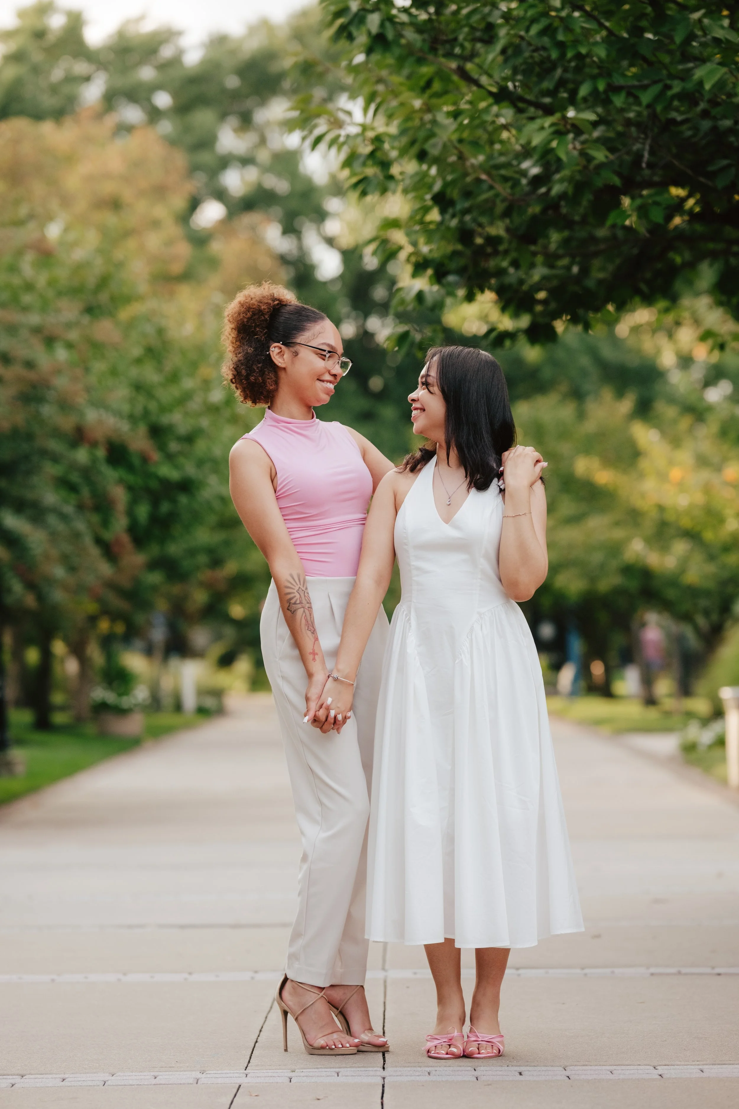 Two women holding hands and smiling at each other in a park, one wearing a pink sleeveless top and white pants, and the other wearing a white sleeveless dress.