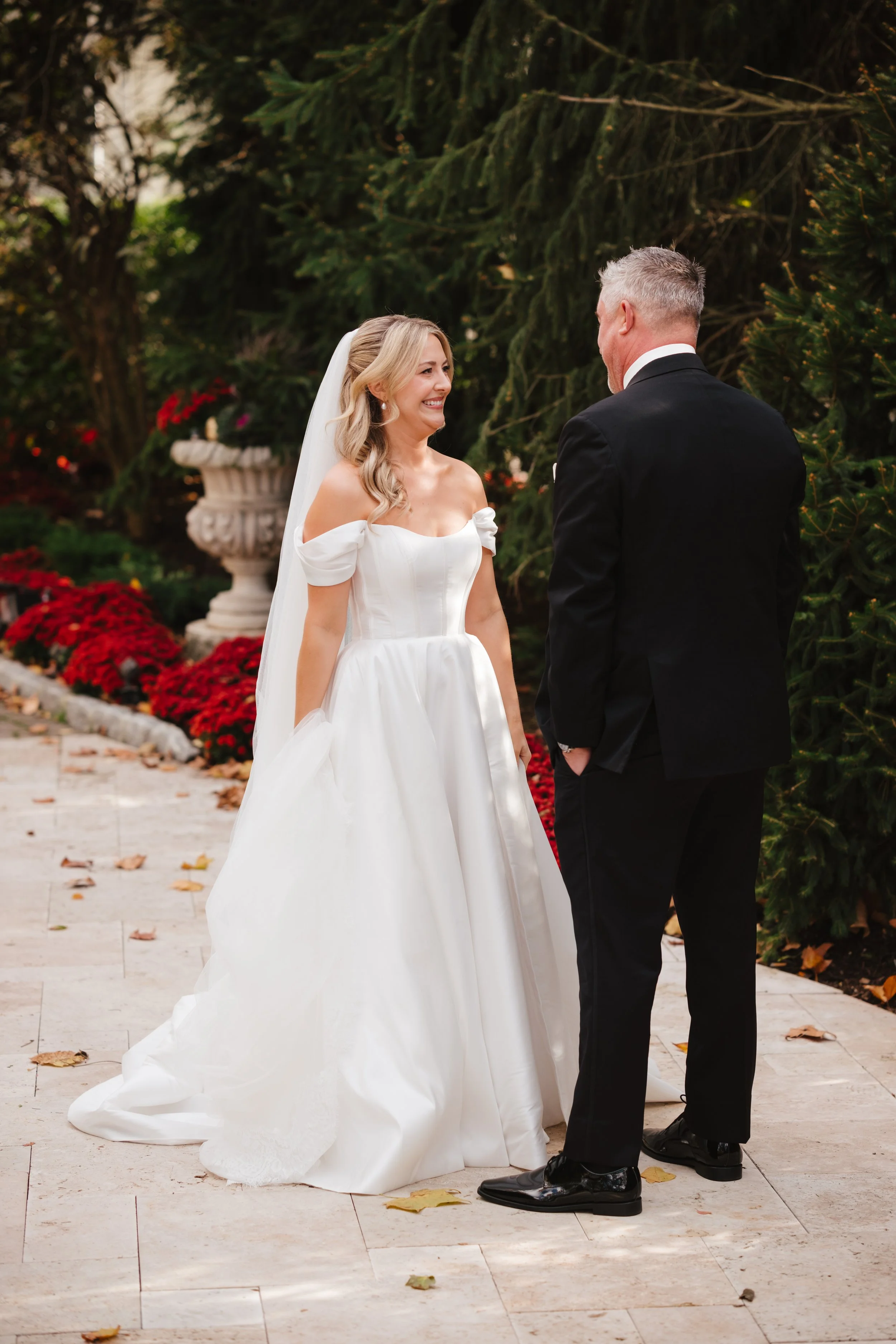 A bride in a white off-shoulder wedding gown and veil is smiling at a groom in a black suit, during an outdoor wedding ceremony with trees and red flowers in the background.