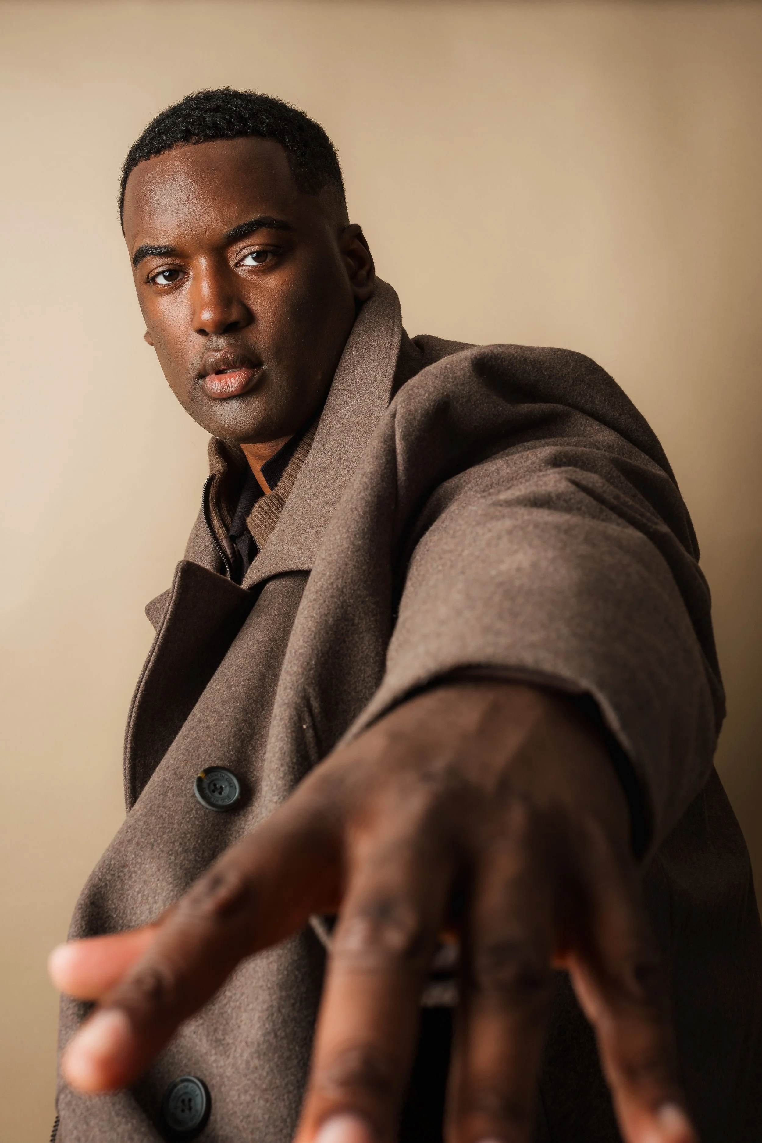 A young man with short black hair and dark skin points toward the camera, wearing a brown coat over a dark shirt, standing against a plain beige background.