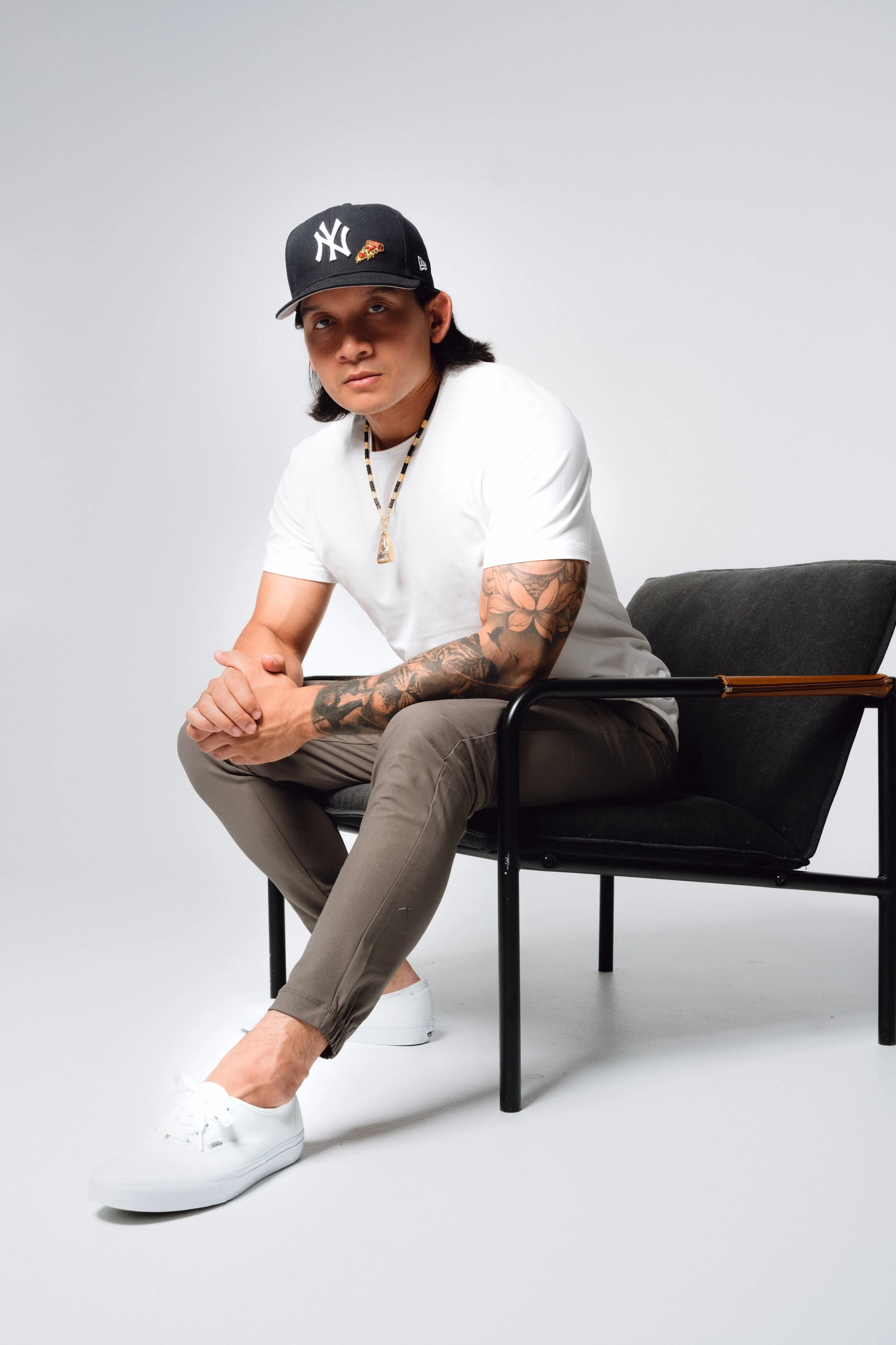 A young man with long dark hair sitting on a black chair, wearing a white t-shirt, beige pants, white sneakers, a black baseball cap with a logo and an embroidered pizza slice, a necklace, and tattoos on his right arm, against a plain white backgroun
