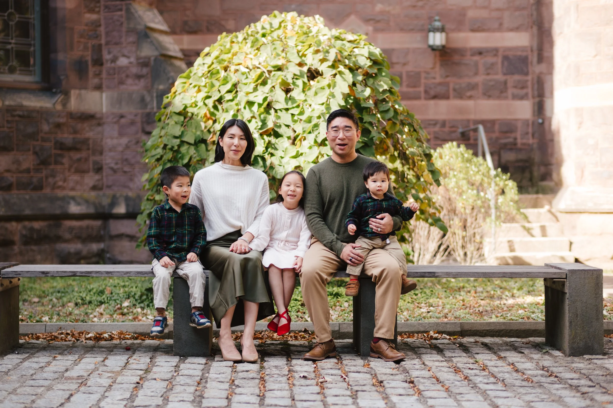 Family of five sitting on a bench outdoors in front of a bush with yellow leaves, with a brick building in the background. The group includes two adults and three children.