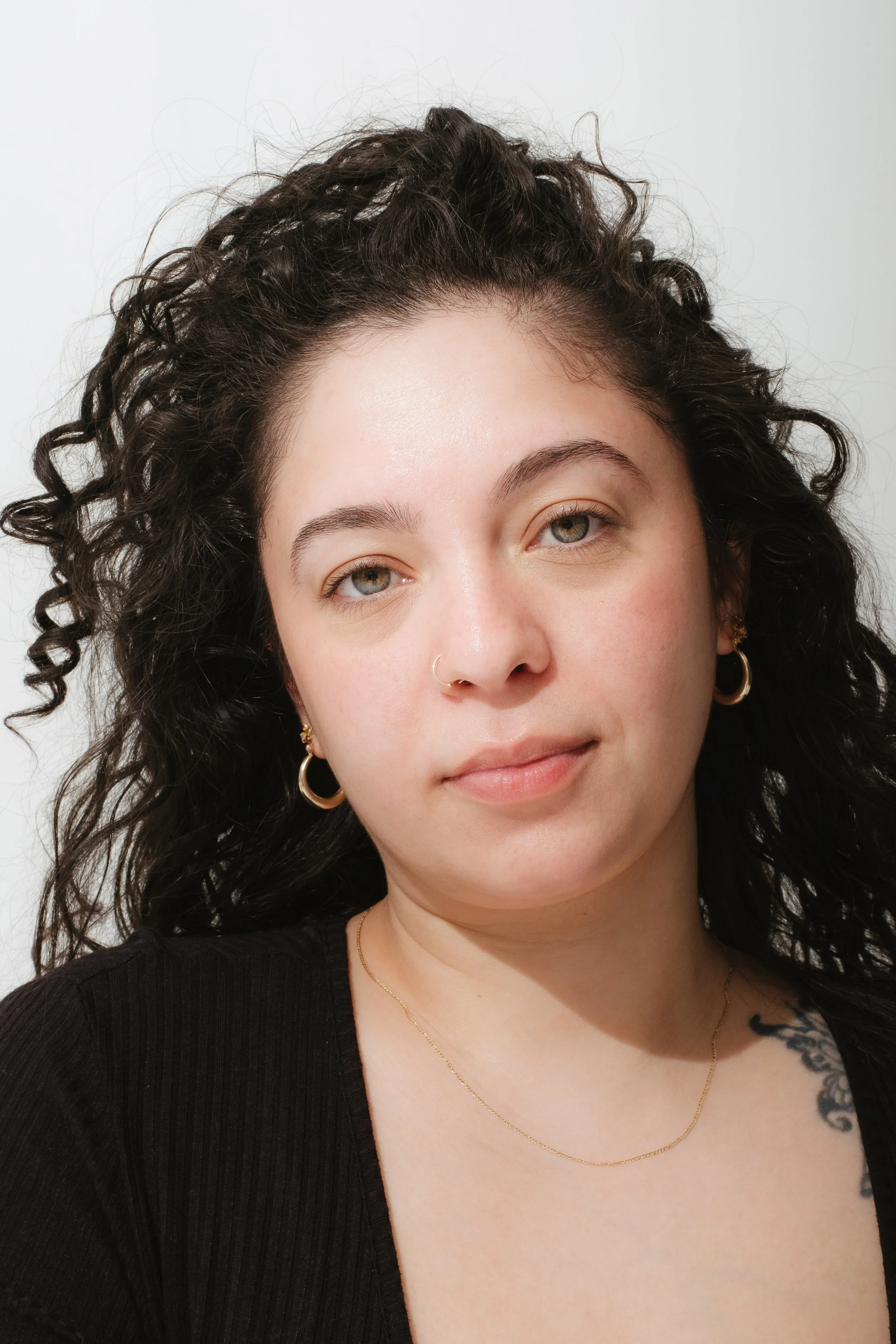 Close-up portrait of a young woman with curly dark hair, wearing gold hoop earrings, a necklace, and a black top, against a plain white background.