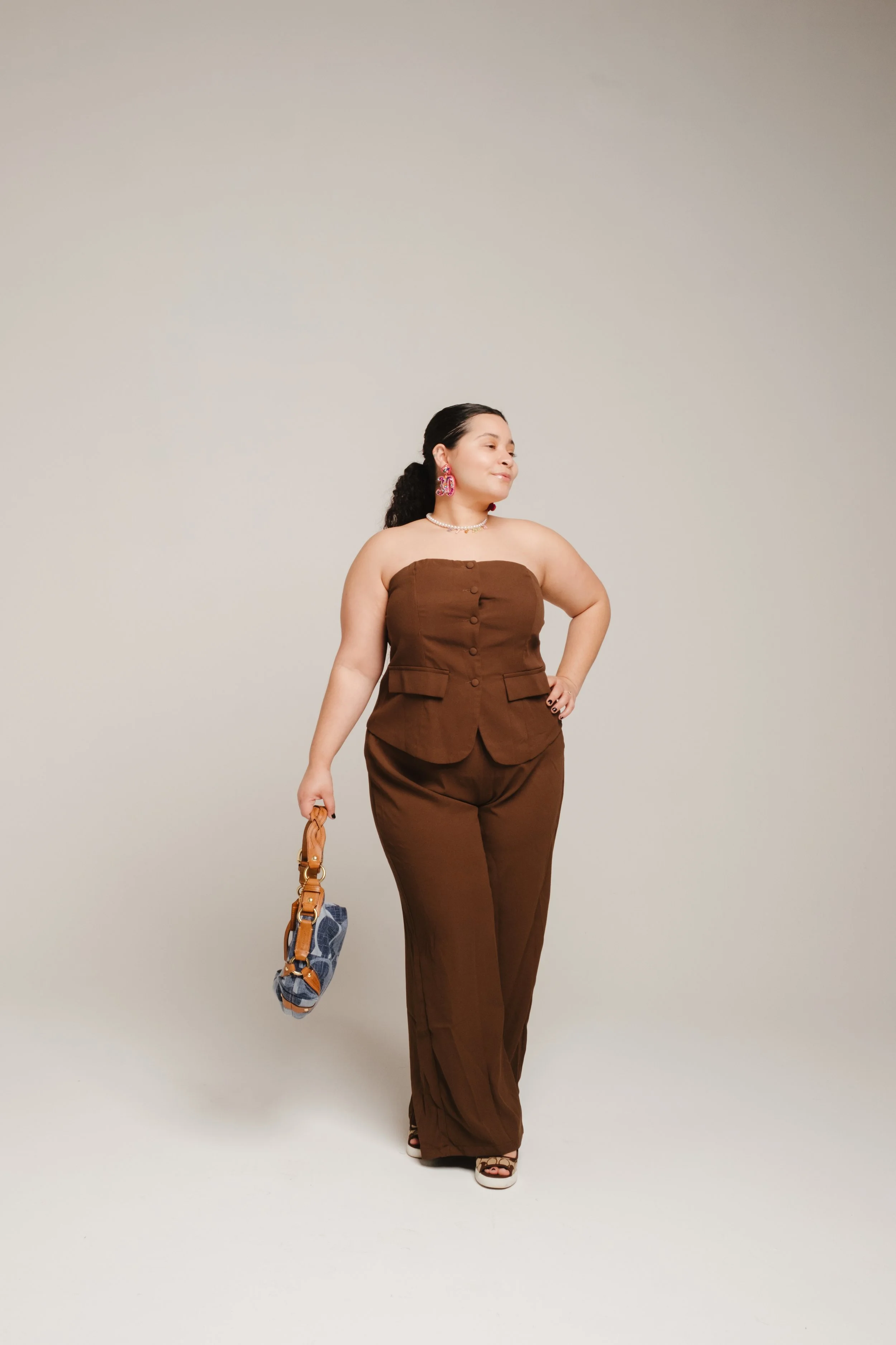 A confident woman in brown trousers and a strapless top posing with her hand on her hip, holding a small handbag, against a plain light gray background.