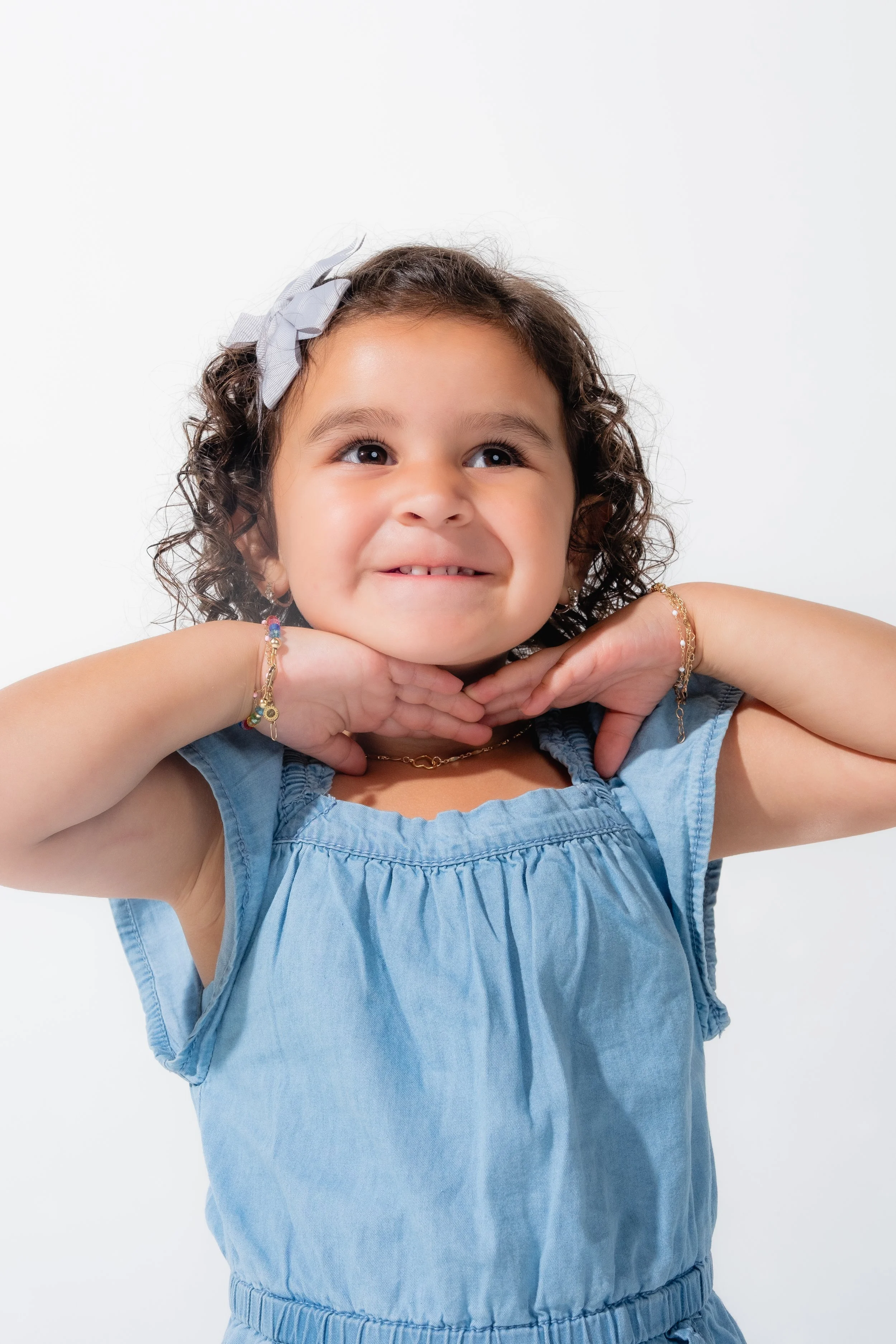 A young girl with curly hair wearing a light blue dress, smiling with her hands under her chin, against a plain white background.