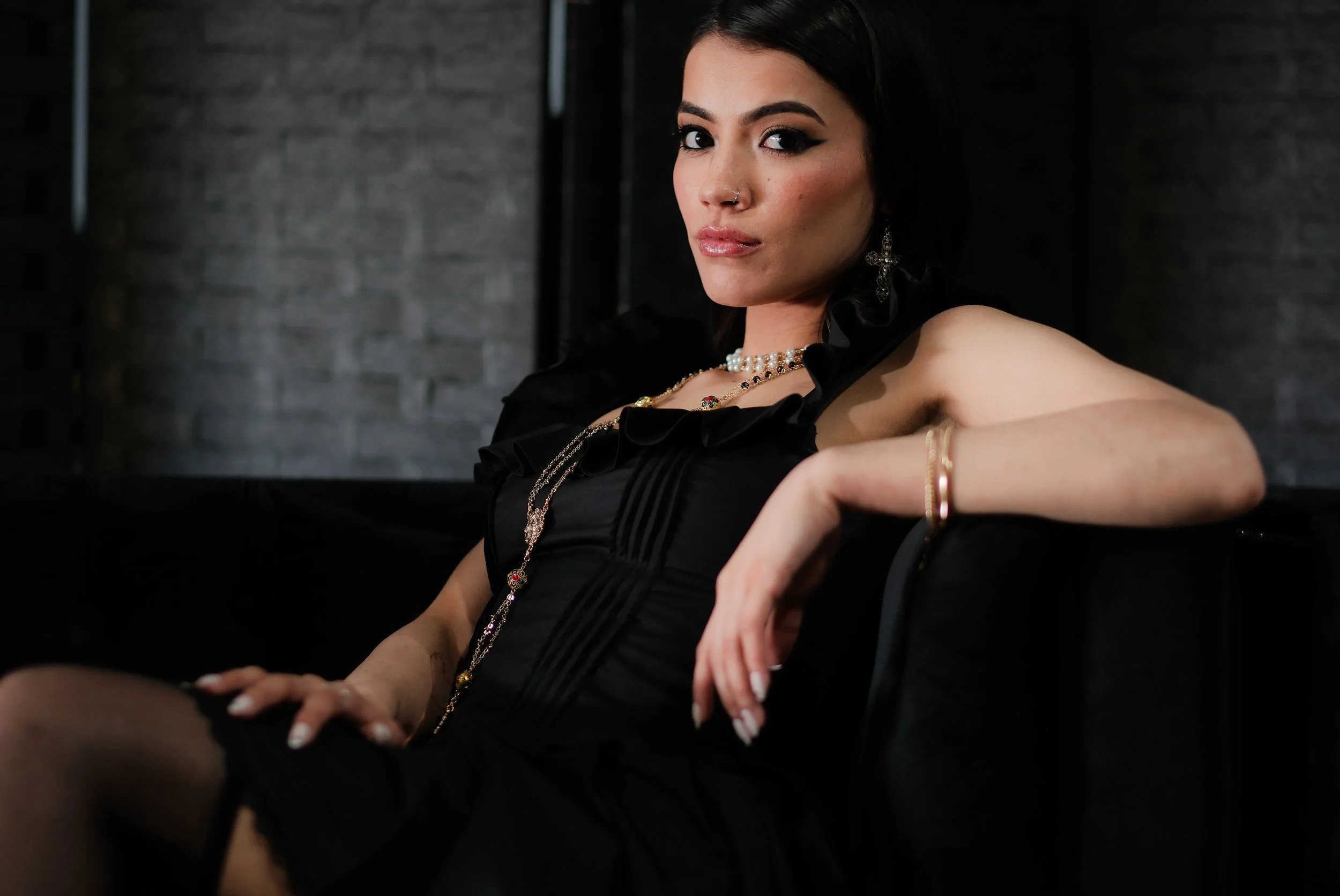 A woman with dark hair, makeup, and jewelry sitting on a black sofa against a dark brick wall.