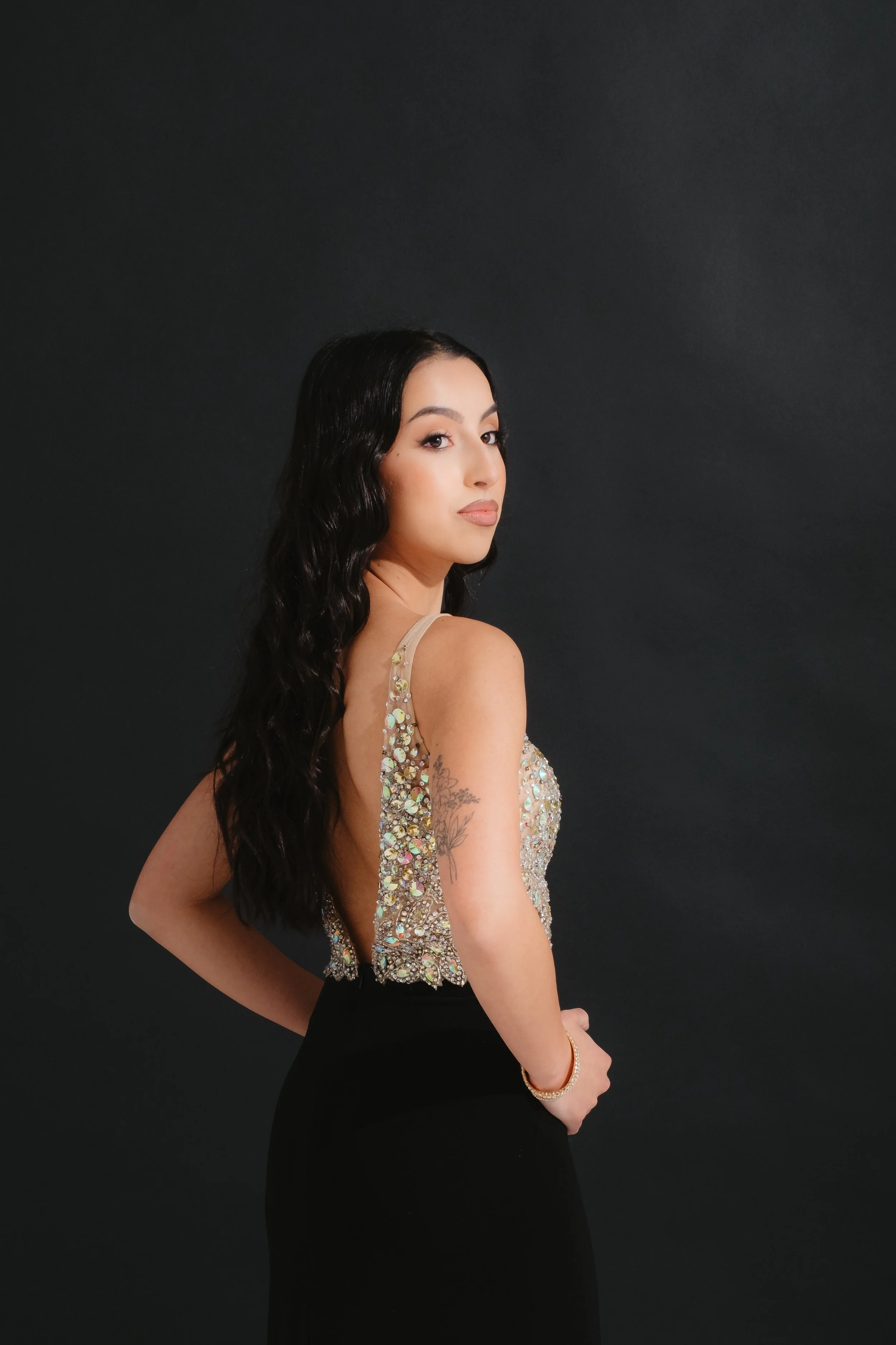 A woman with long black wavy hair, wearing a sparkling, embellished top and a black skirt, stands against a black background, posing with her body turned to the side and her head facing forward.