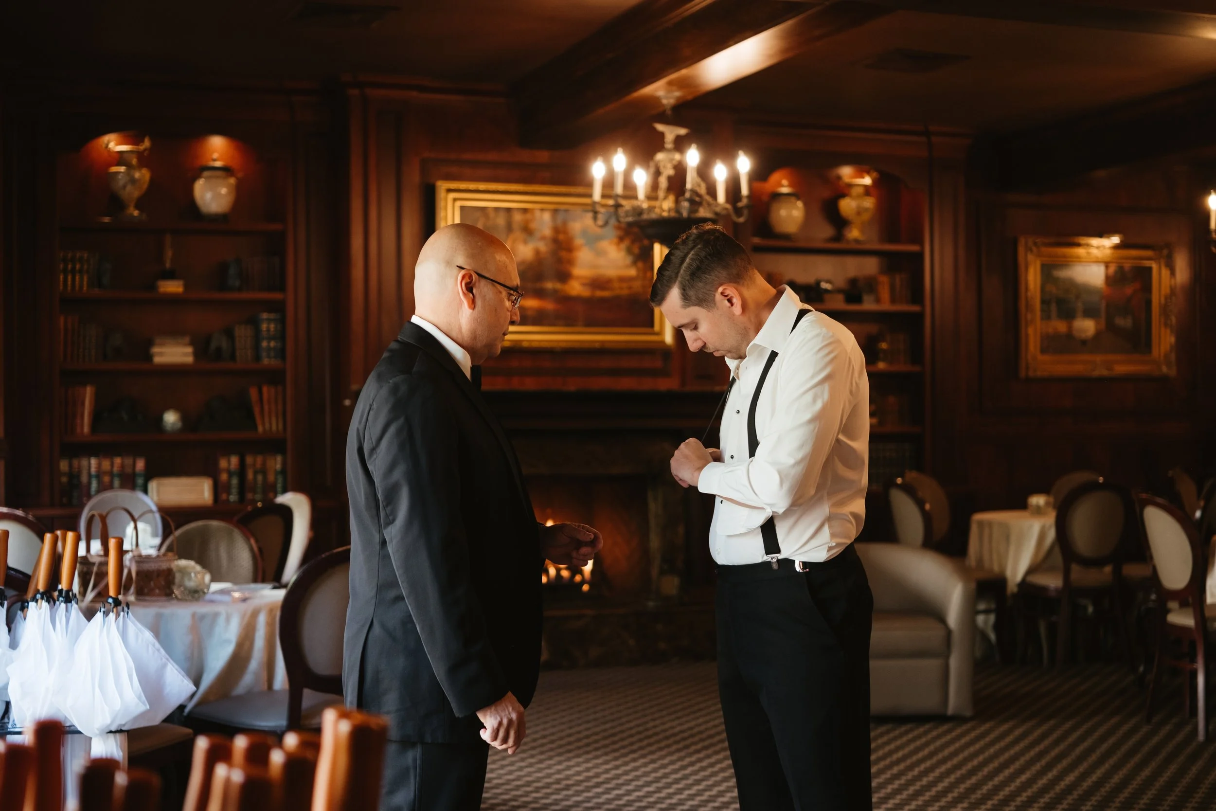 Two men in formal attire standing in a dimly lit, wood-paneled room decorated with paintings, bookshelves, and chandeliers, with tables and chairs in the background.