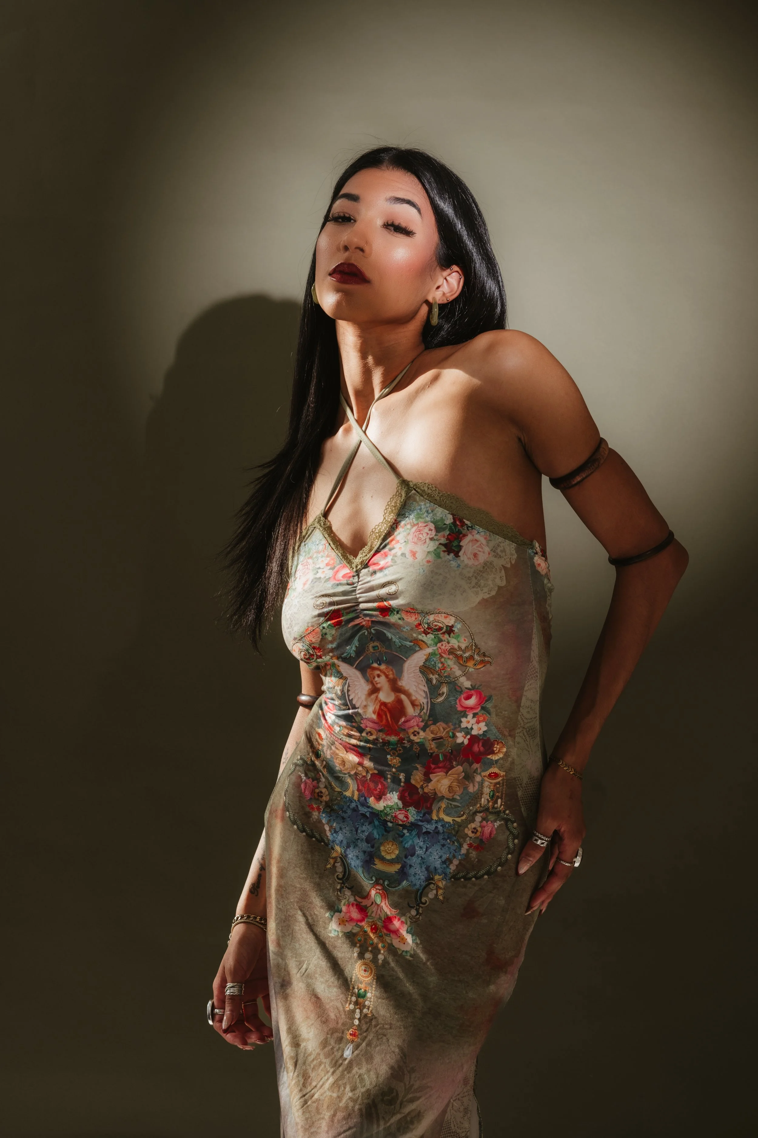 A woman with long dark hair and bold makeup wearing a colorful, floral printed dress with a halter neck, posing against a plain wall.