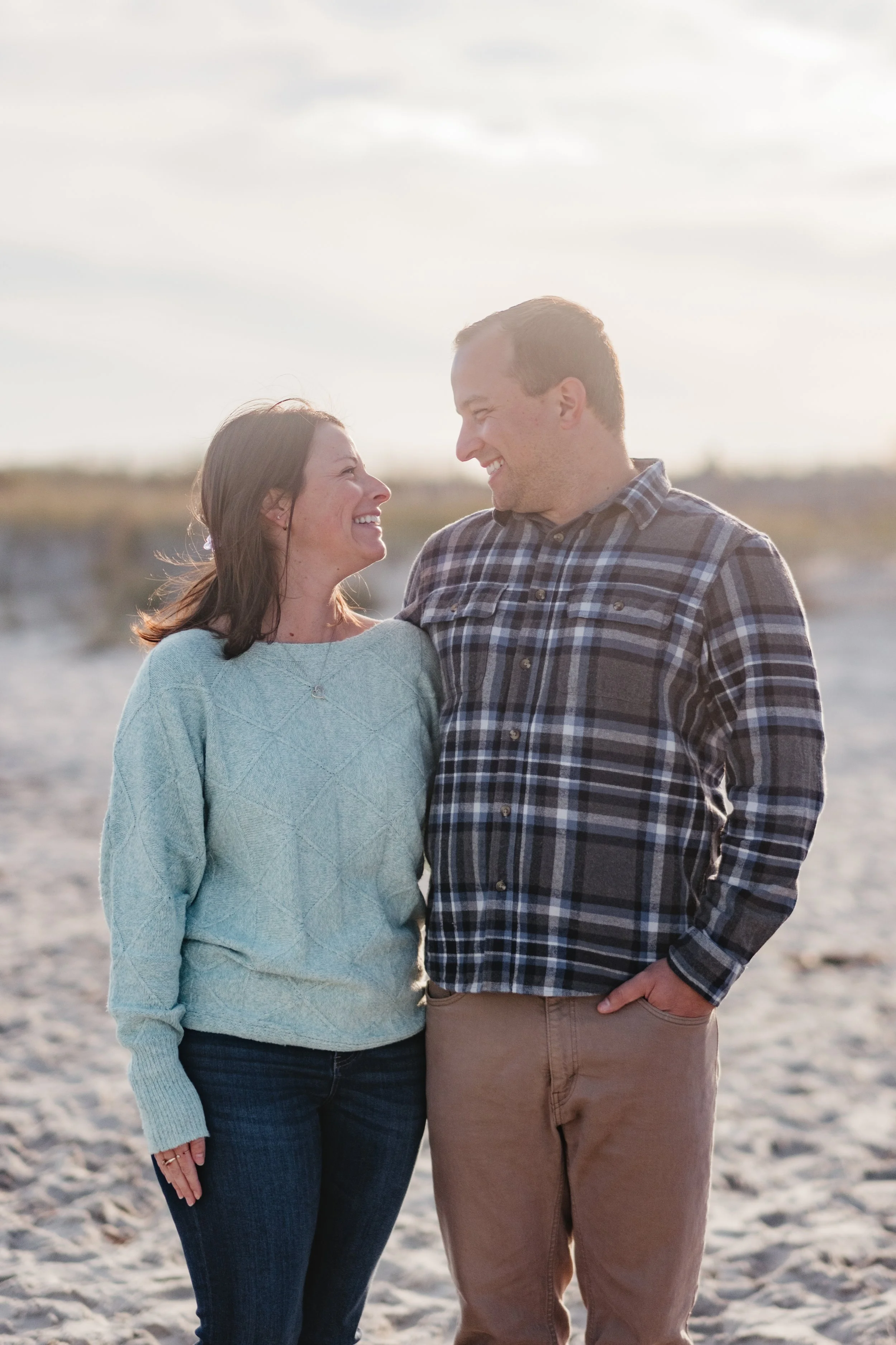 A smiling couple, a woman in a light blue sweater and a man in a plaid shirt, gazing at each other on a beach during sunset.