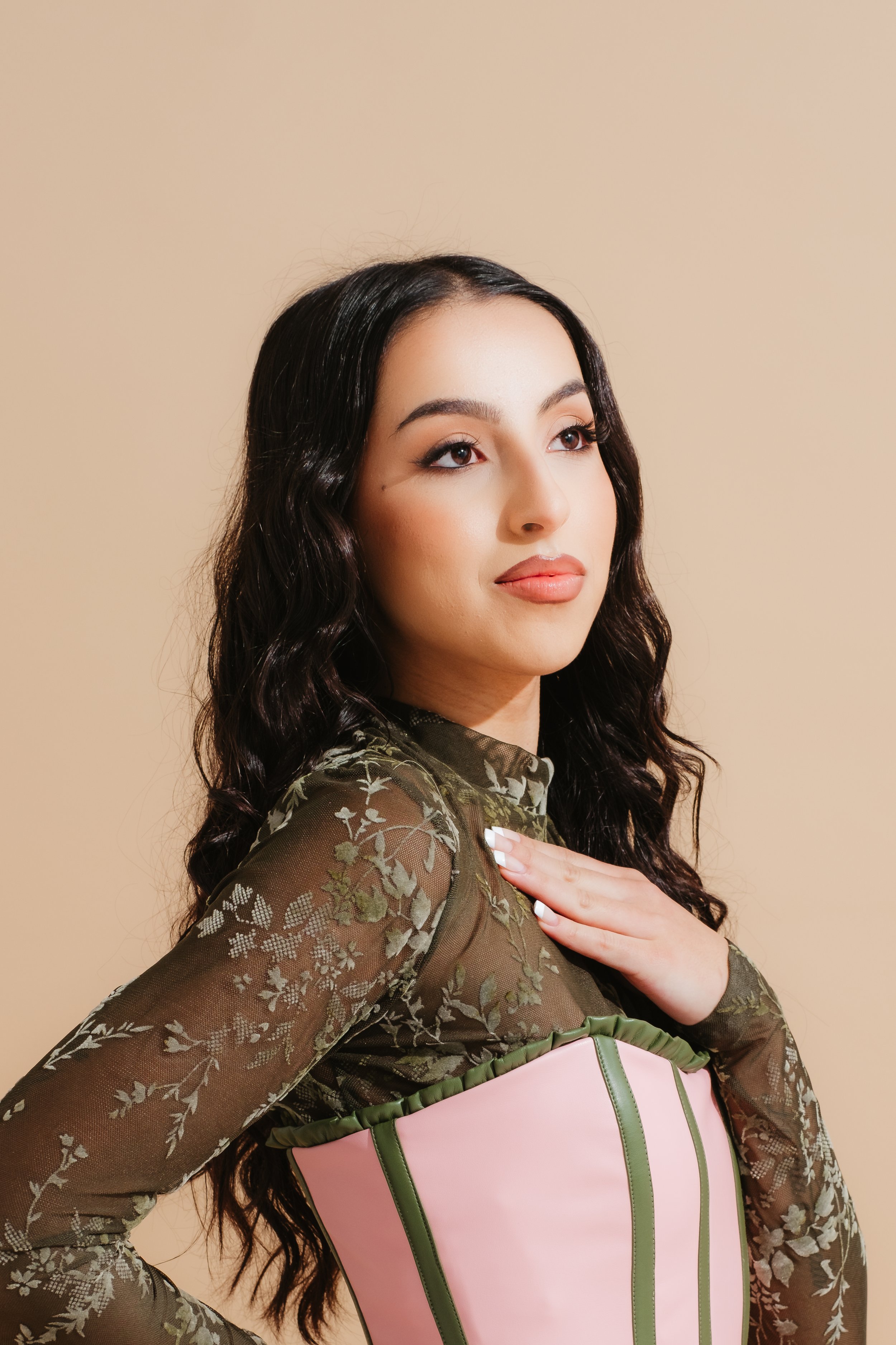 A young woman with dark wavy hair wearing a sheer, floral-patterned top and holding a pink bag with green accents over her arm, standing against a beige background.