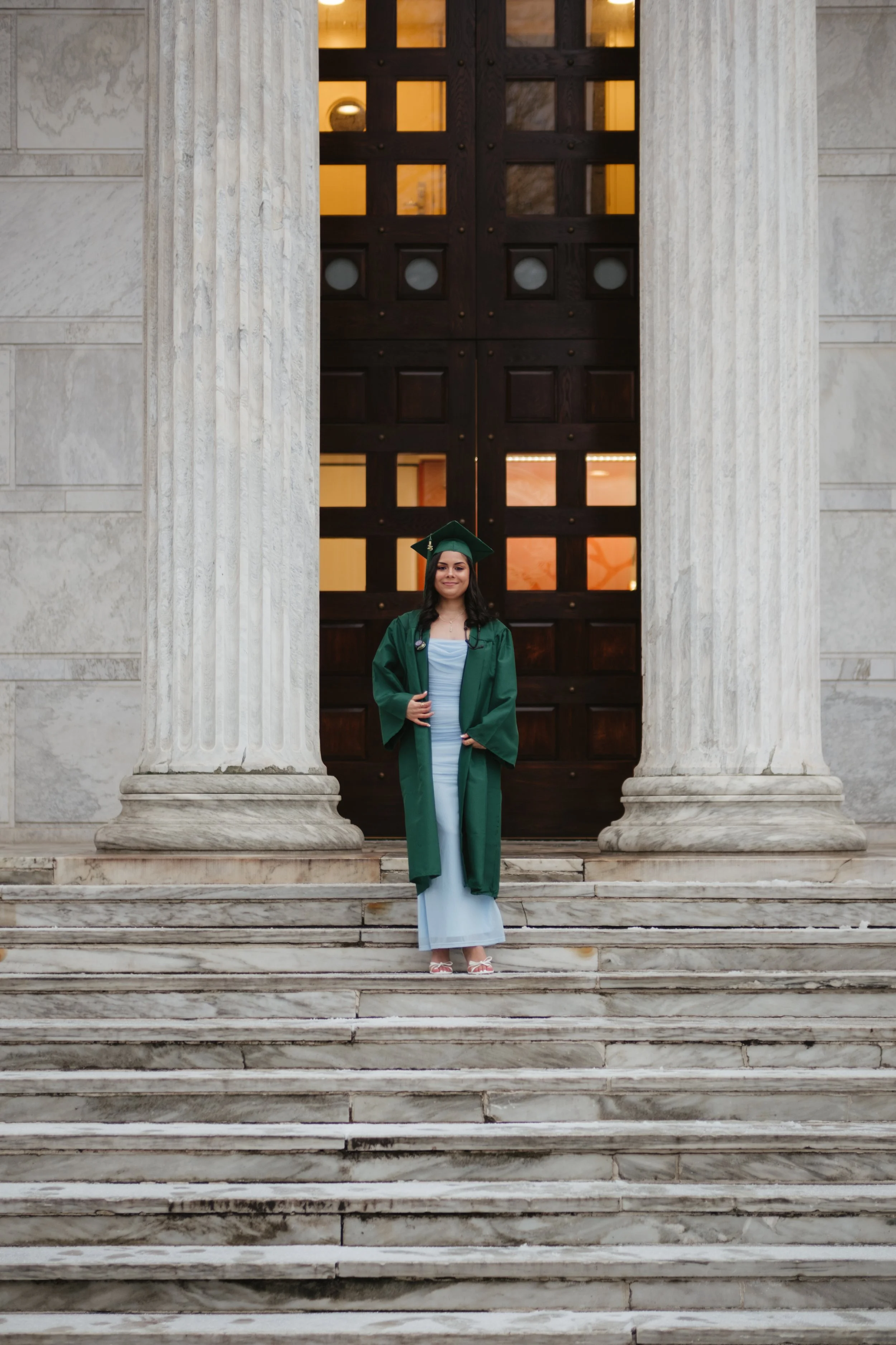 A young woman in a green graduation gown and cap standing on marble steps in front of large columns and a wooden door, smiling.