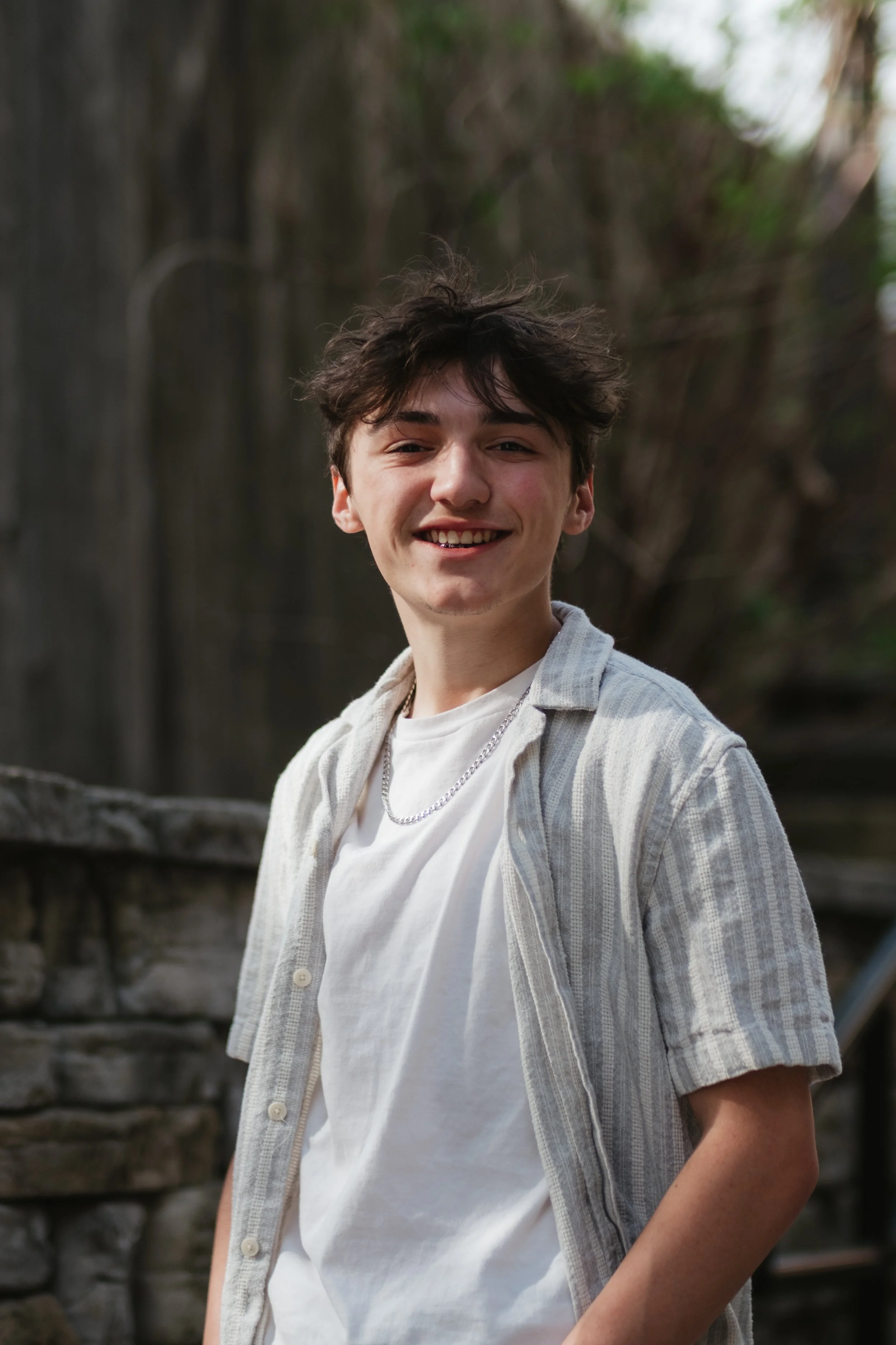 A young man with dark, messy hair is smiling outdoors in front of a wooden fence and greenery, wearing a white t-shirt, an open striped shirt, and a silver chain necklace.