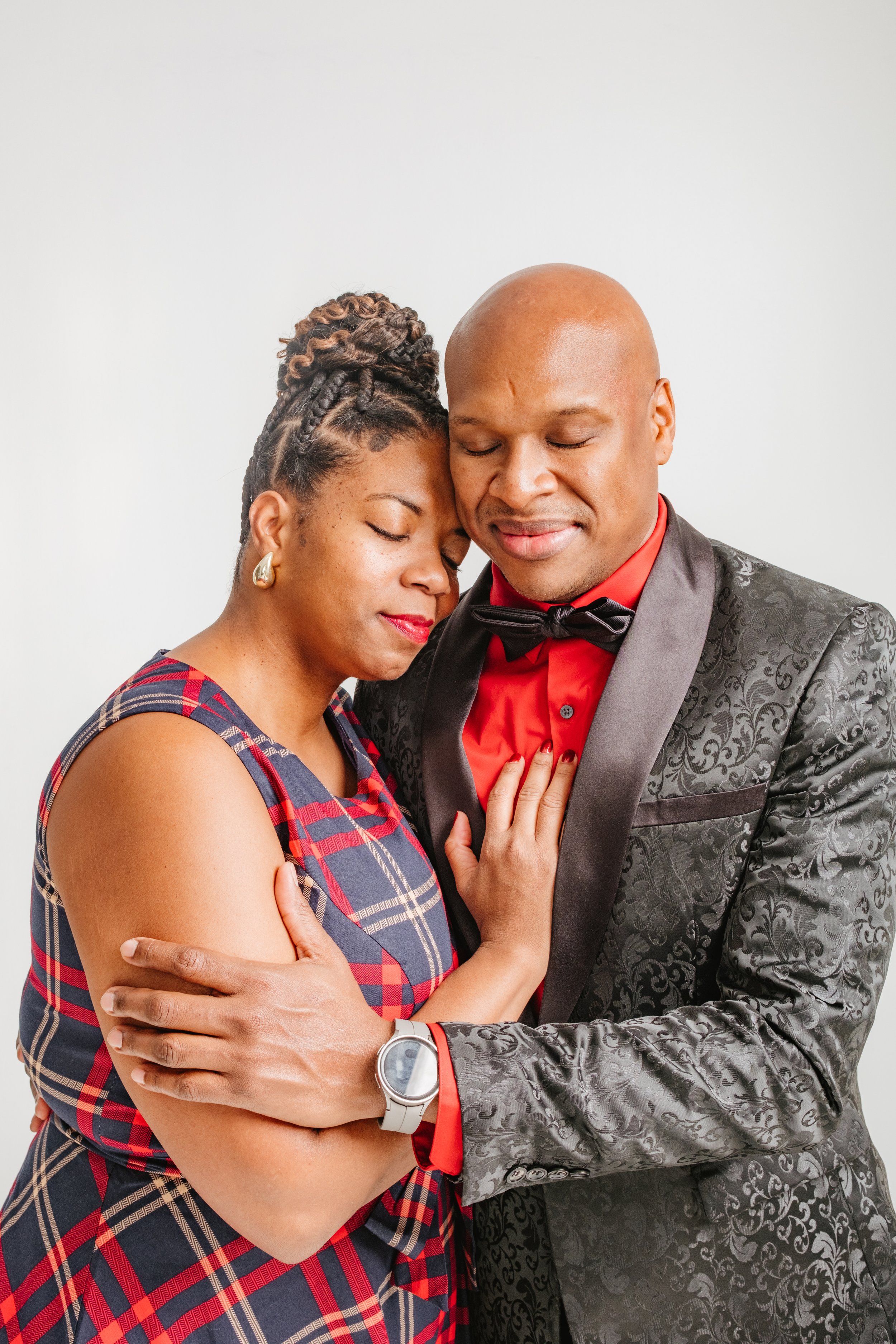 A couple dressed in formal attire embracing with eyes closed and smiling, standing against a plain white background.