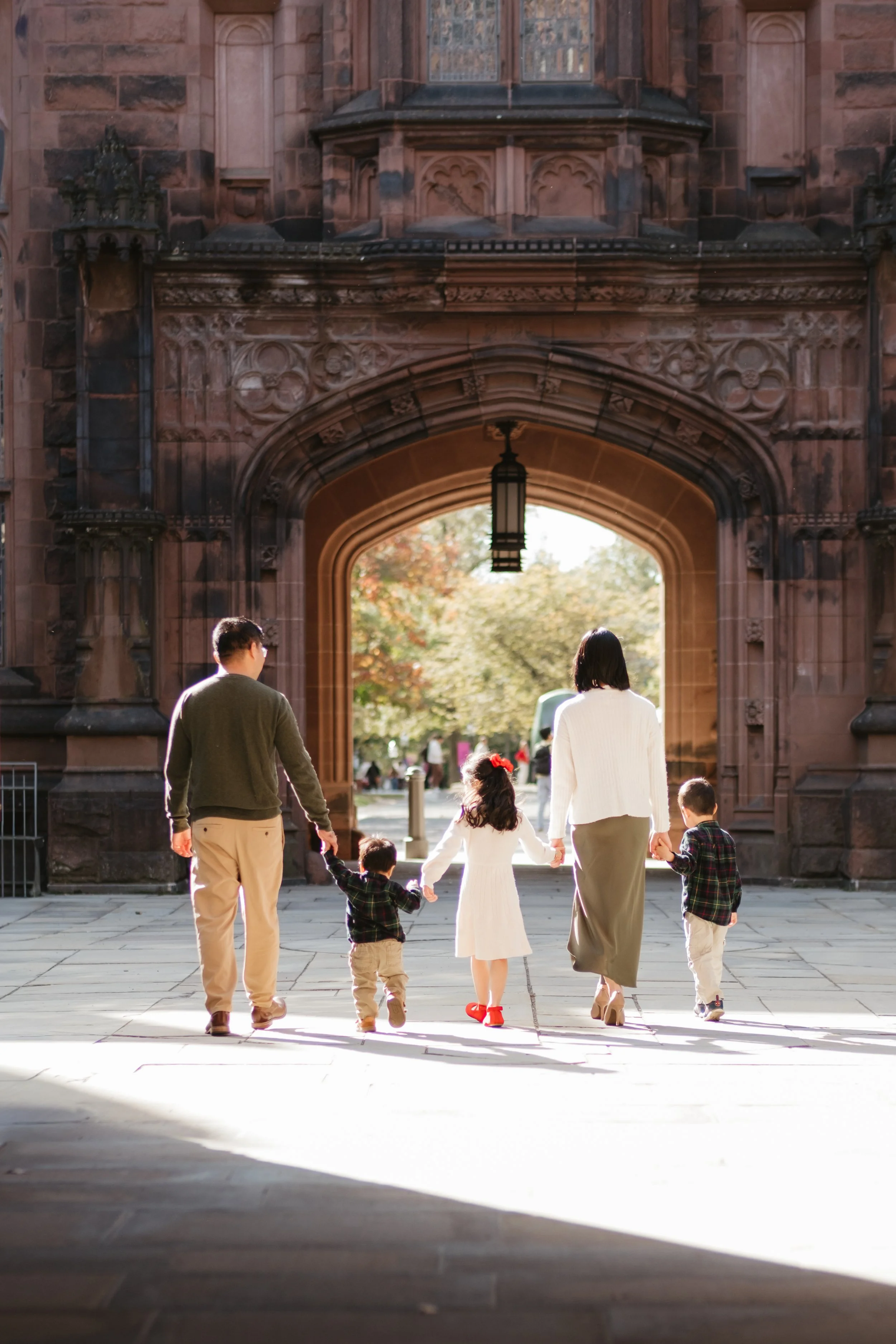 A family of five walking hand in hand through an arched stone gateway on a sunny day, with trees in the background.