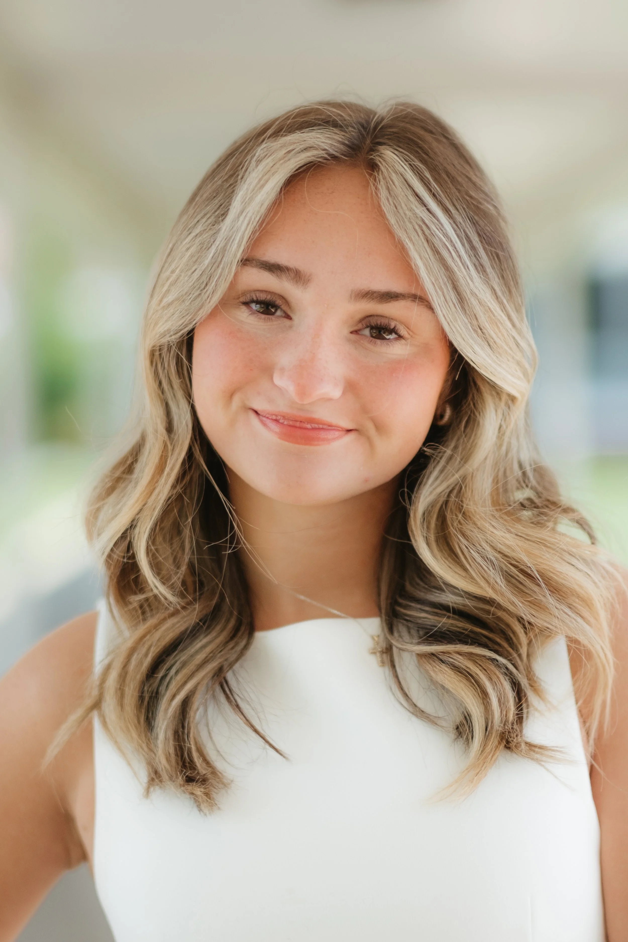 Young woman with shoulder-length wavy blonde hair, wearing a white sleeveless top and a gold cross necklace, smiling outdoors.