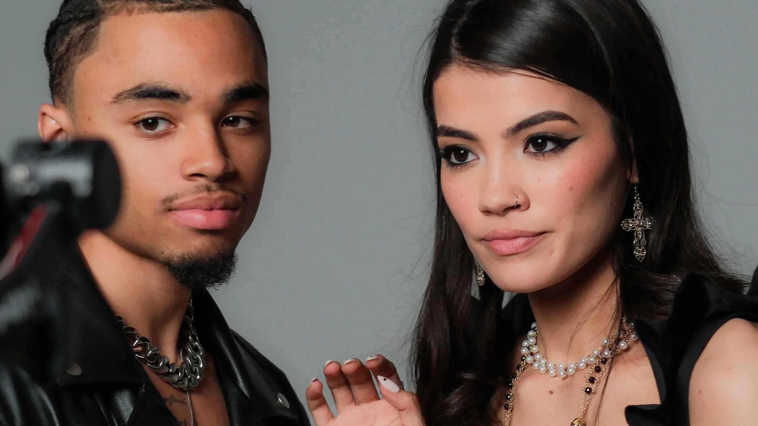 Two young adults, a man and a woman, are posing closely for a photo against a plain background. The man has short, styled hair, and is wearing a black leather jacket with a chain necklace. The woman has long dark hair, makeup with winged eyeliner, la