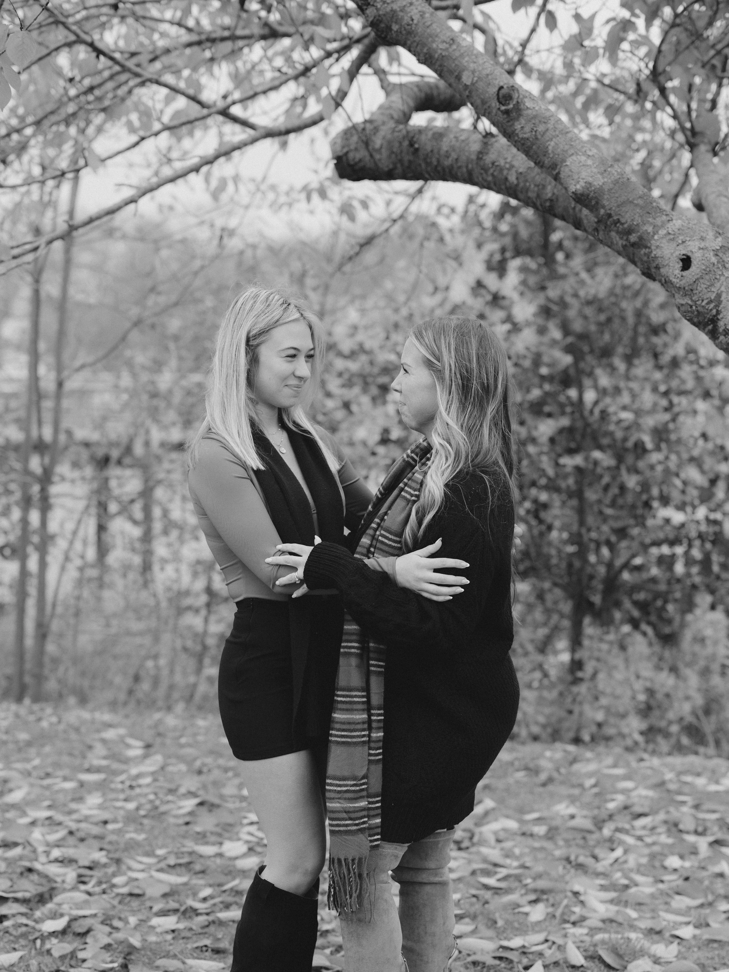 Two women standing close together outdoors under a tree, looking at each other and smiling, with fallen leaves on the ground.