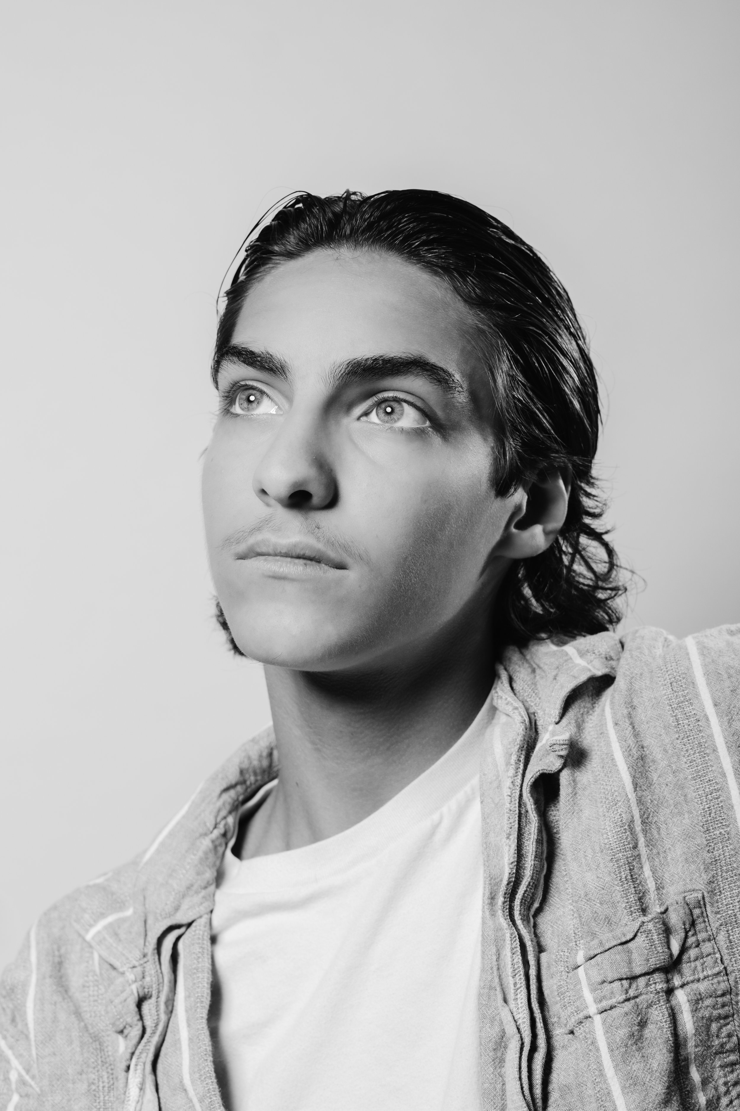 Black and white portrait of a young man with slicked-back hair, wearing a casual jacket over a white t-shirt, looking slightly upwards.
