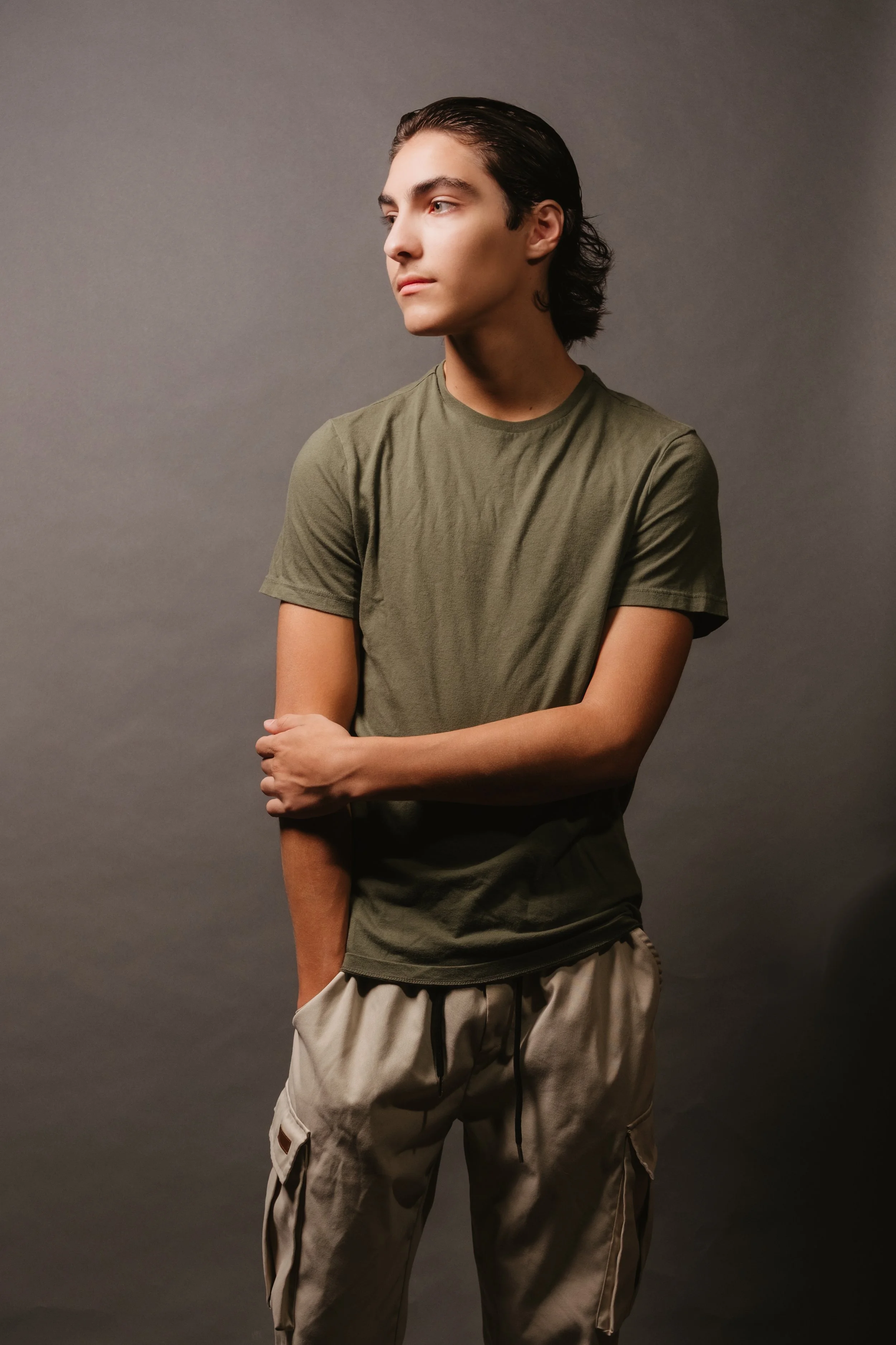 A young man with dark, slicked-back hair, wearing a green t-shirt and beige cargo pants, is standing against a plain gray background, looking away to his right with his left arm crossed over his waist and his right arm resting on it.