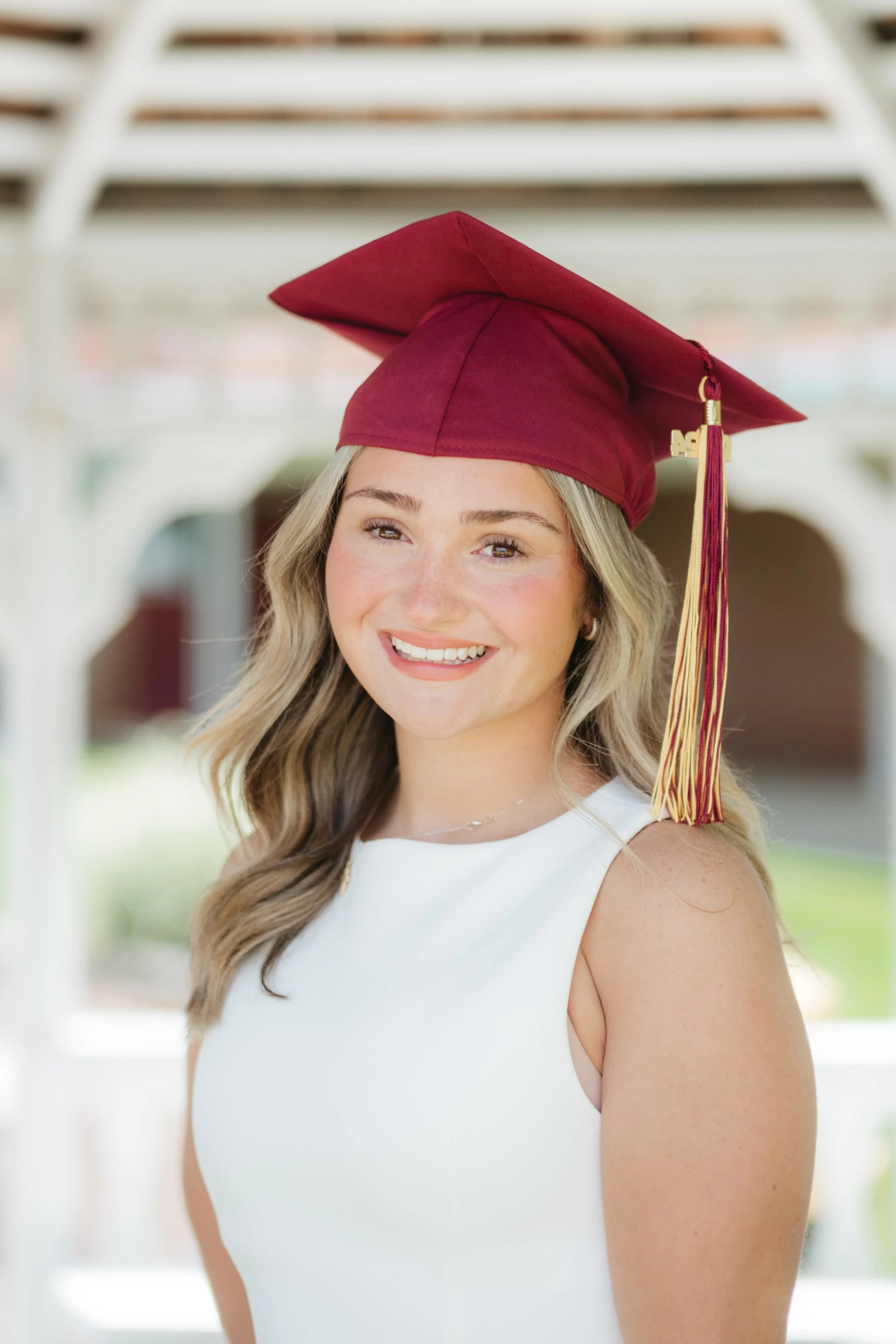 A young woman wearing a white sleeveless dress and a maroon graduation cap with a gold and maroon tassel, smiling outdoors.