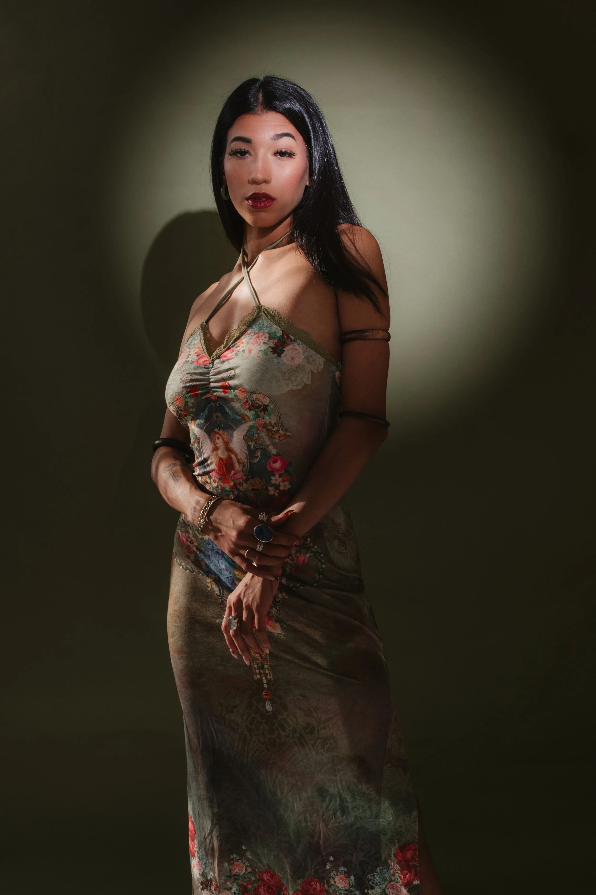 A woman with black hair and red lipstick standing against a plain, olive-green background, wearing a sleeveless, floral and patterned dress with accessories including rings, bracelets, and arm bands.