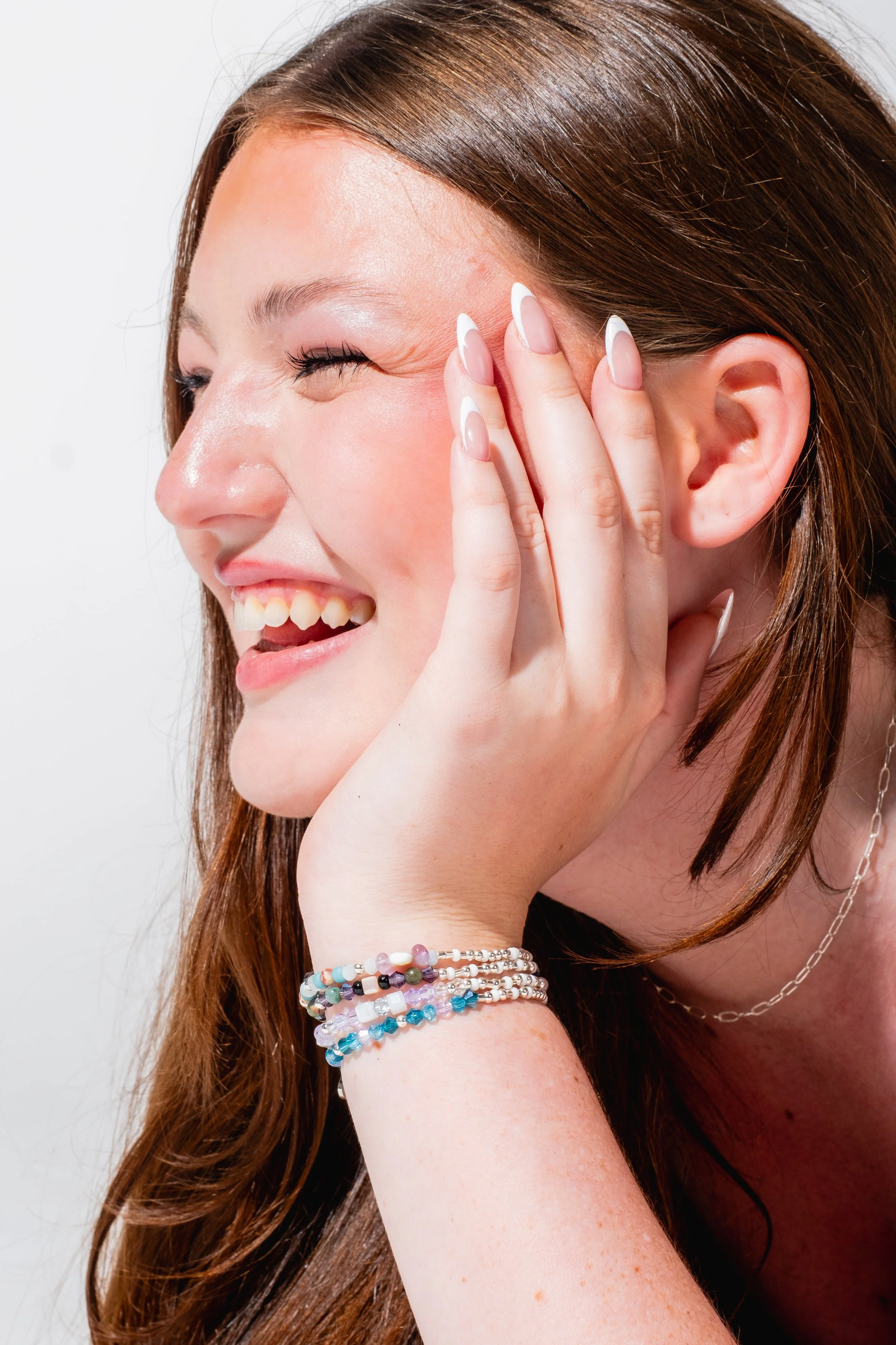 A woman with long brown hair smiling and laughing, touching her cheek with her hand, wearing multiple beaded bracelets and a thin chain necklace.