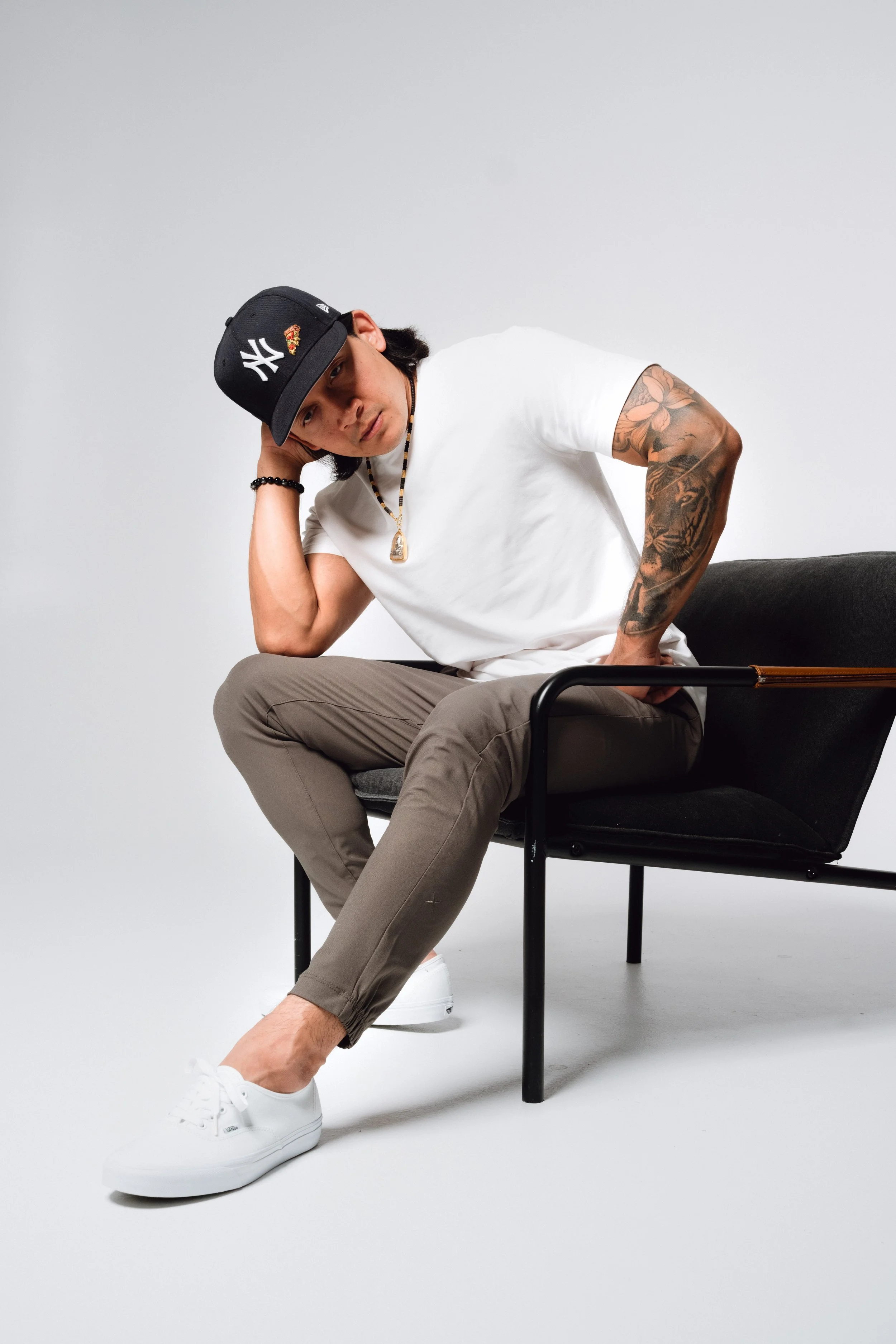 Young man sitting on a black chair in front of a plain white background, wearing a black New York Yankees cap, white t-shirt, beige pants, and white sneakers, with tattoos on his left arm, a beaded bracelet, a necklace, and a hand resting on his head