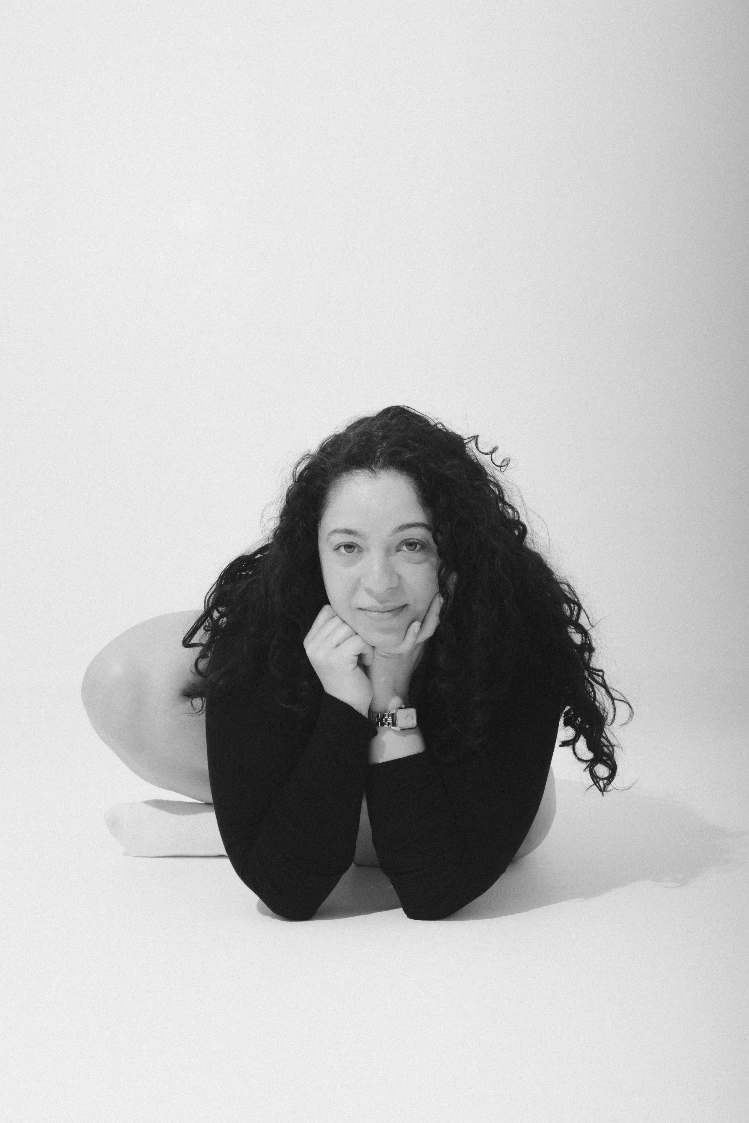 A woman lying on her stomach on a white surface with her chin resting on her hands, wearing a black long-sleeved top, with curly hair, and a watch on her left wrist, against a plain white background.