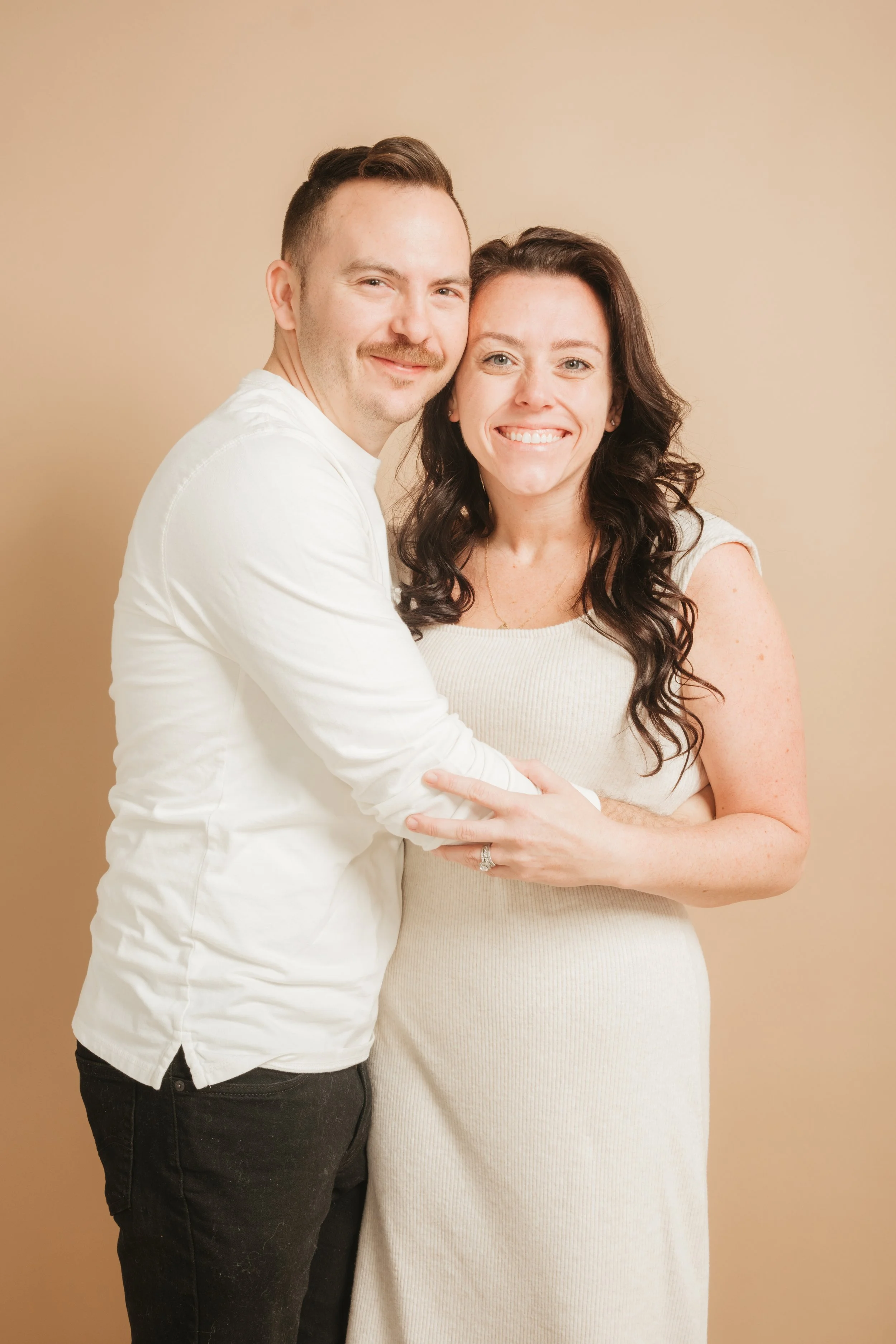 A smiling couple stands together, the man with short brown hair and mustache, the woman with long dark curly hair, both wearing white, embracing each other against a neutral beige background.