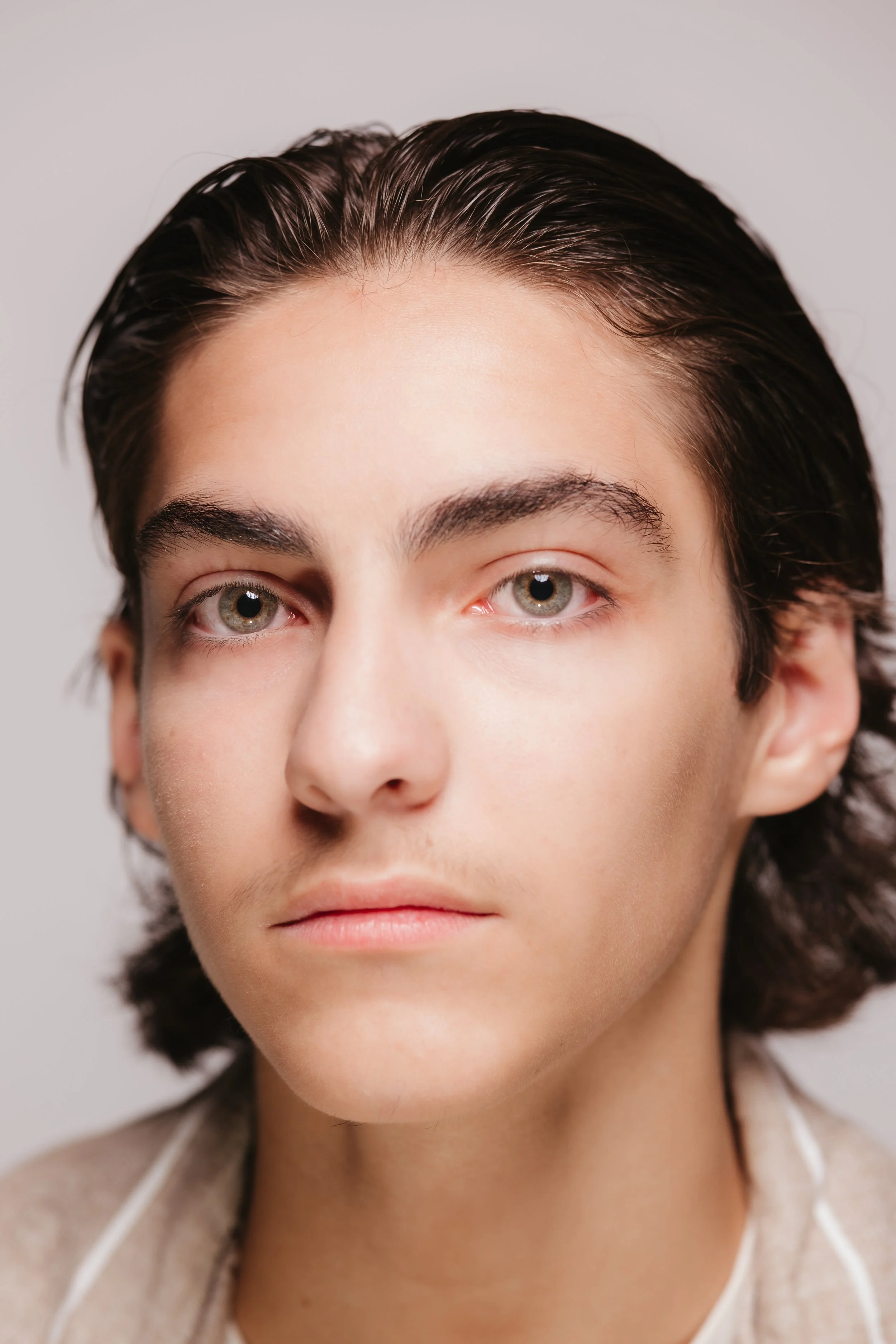Close-up portrait of a young person with dark, wavy hair, fair skin, and light-colored eyes, looking directly at the camera.