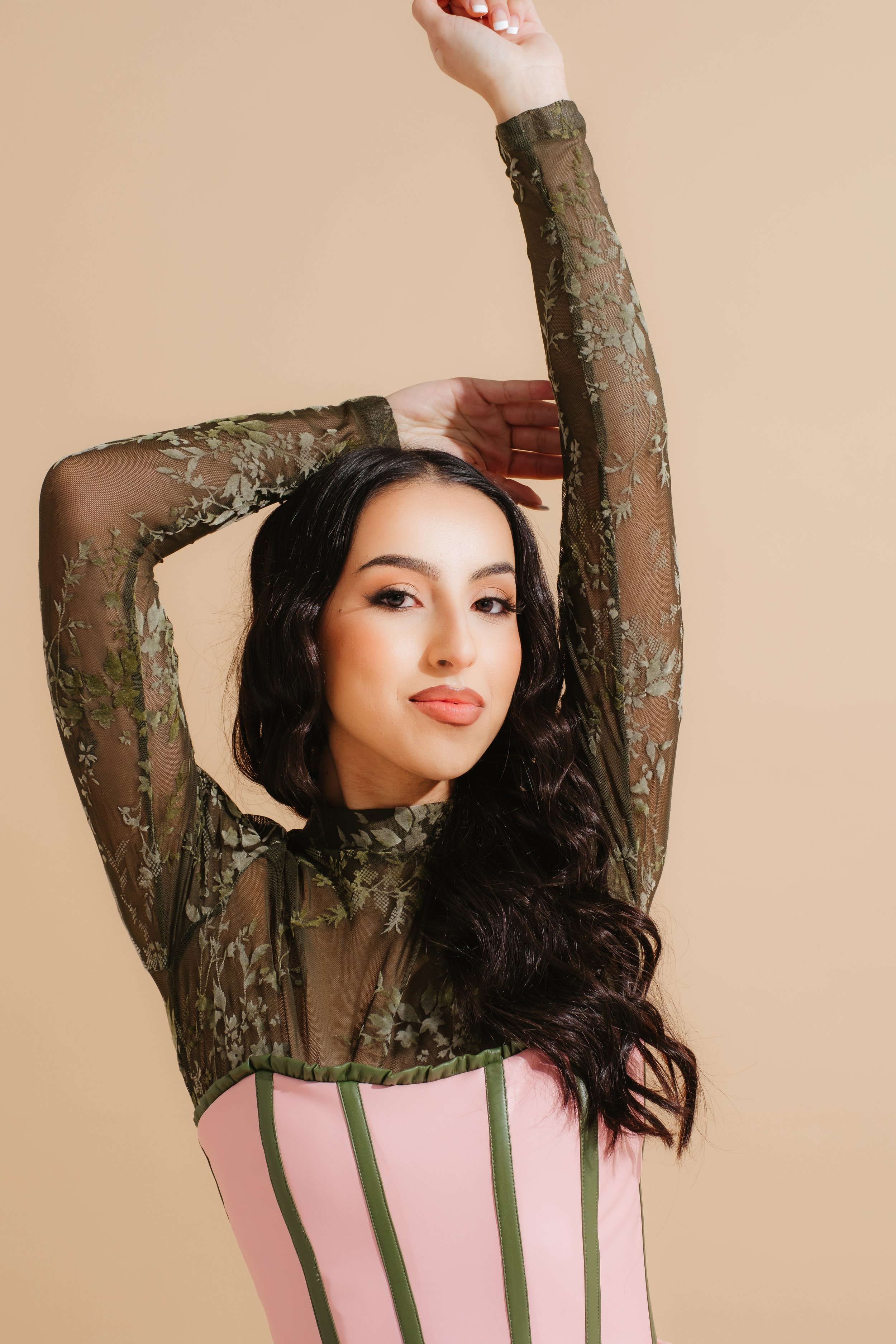 A woman with long dark wavy hair wearing a sheer black floral lace top and a pink corset with green vertical stripes, posing with one arm raised and the other behind her head against a beige background.
