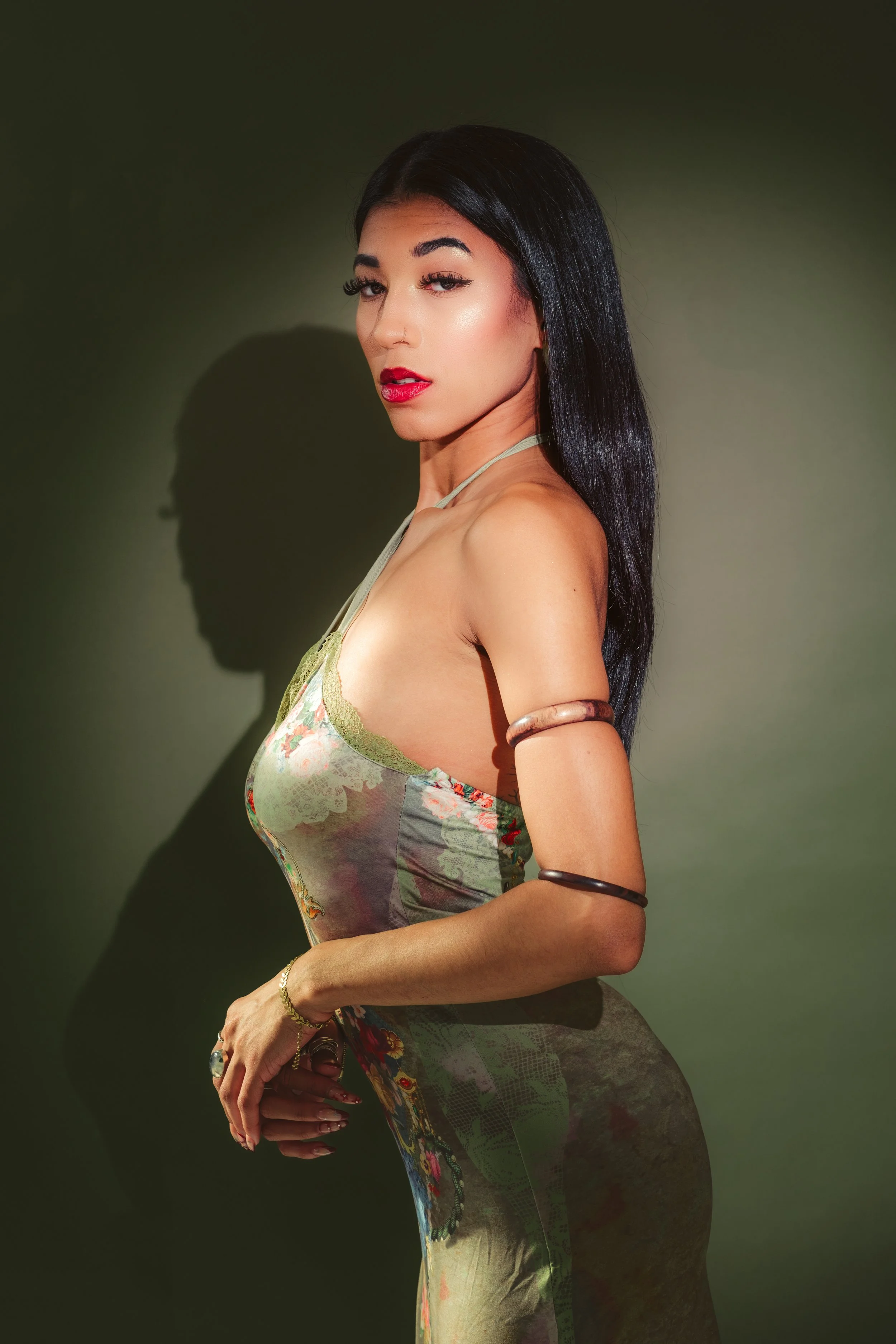 A woman with long black hair, red lipstick, and makeup, wearing a floral dress with straps and jewelry, standing against a dark green background.