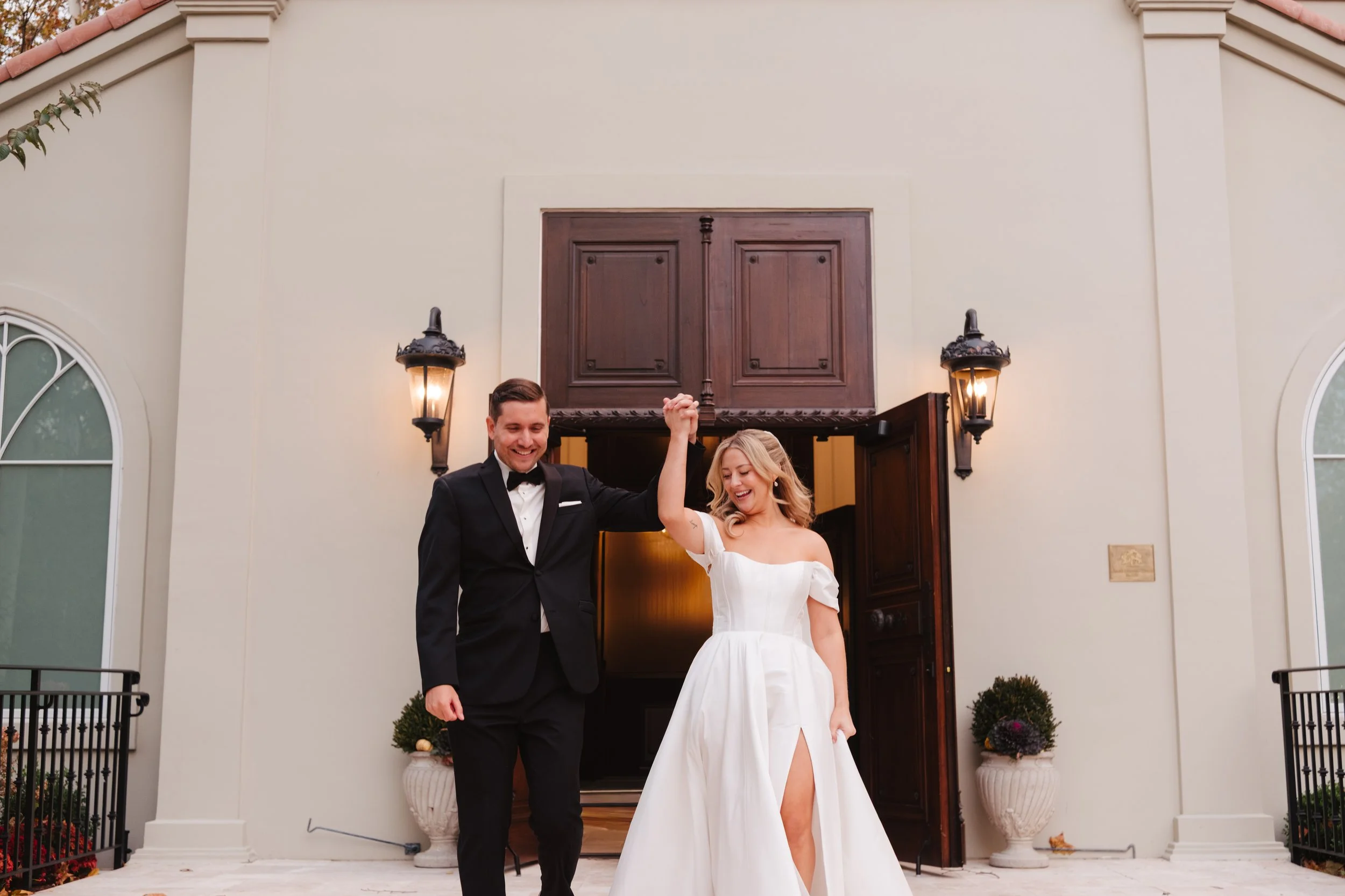 A newlywed couple celebrates outside a building; the groom in a black tuxedo and the bride in a white wedding gown hold hands and smile.