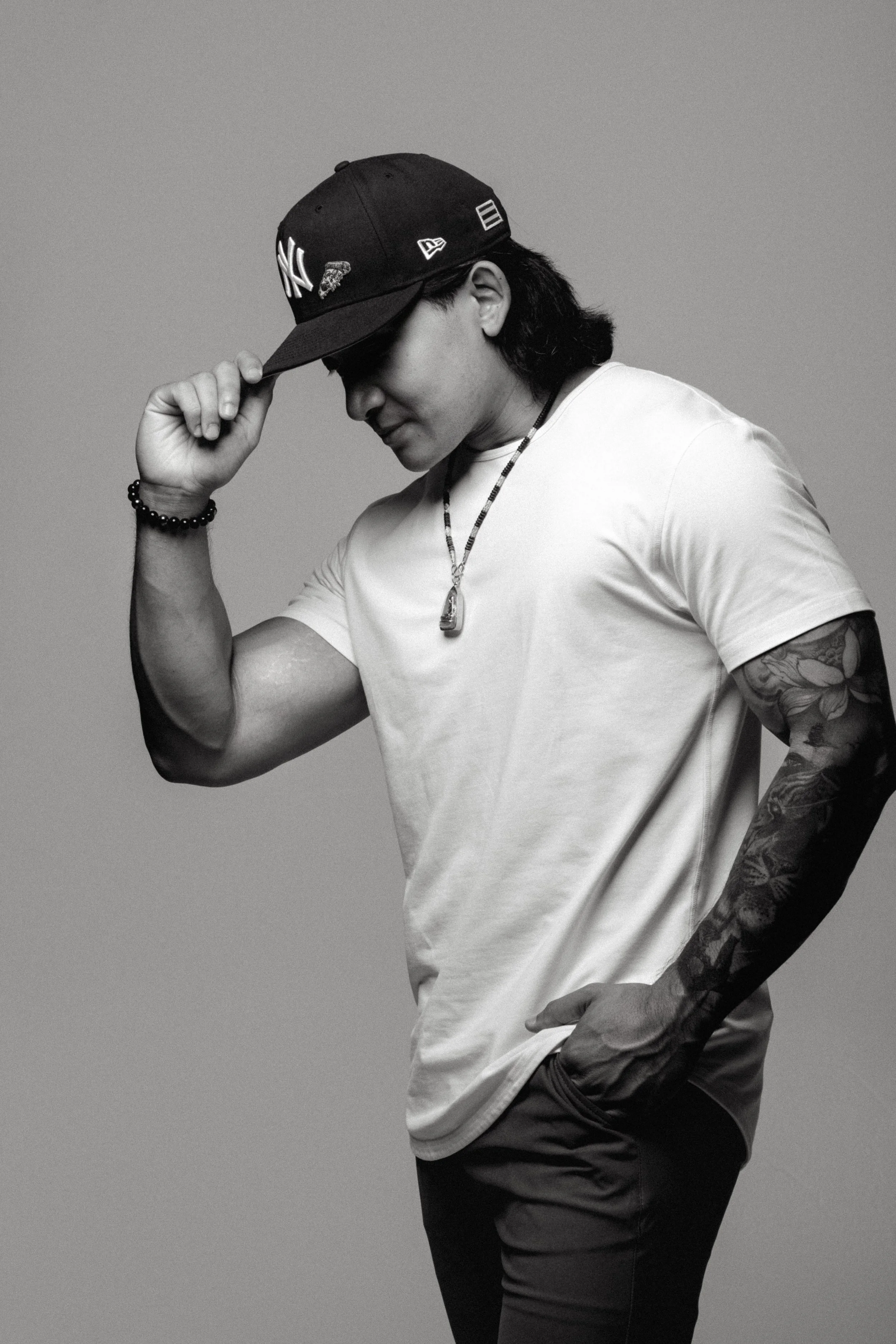 A young man in a white t-shirt and a New York Yankees cap, wearing bracelets and a necklace, posing with his hand on his cap and looking down, against a plain background.