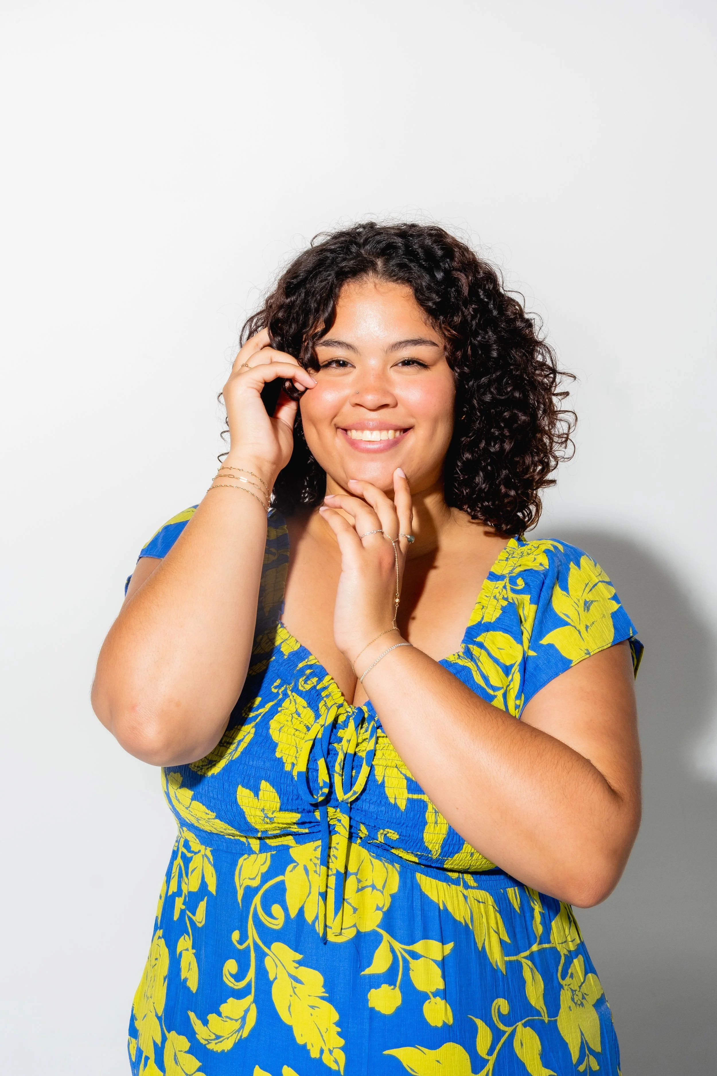 A smiling woman with curly dark hair wearing a blue and yellow floral dress, standing against a white background.