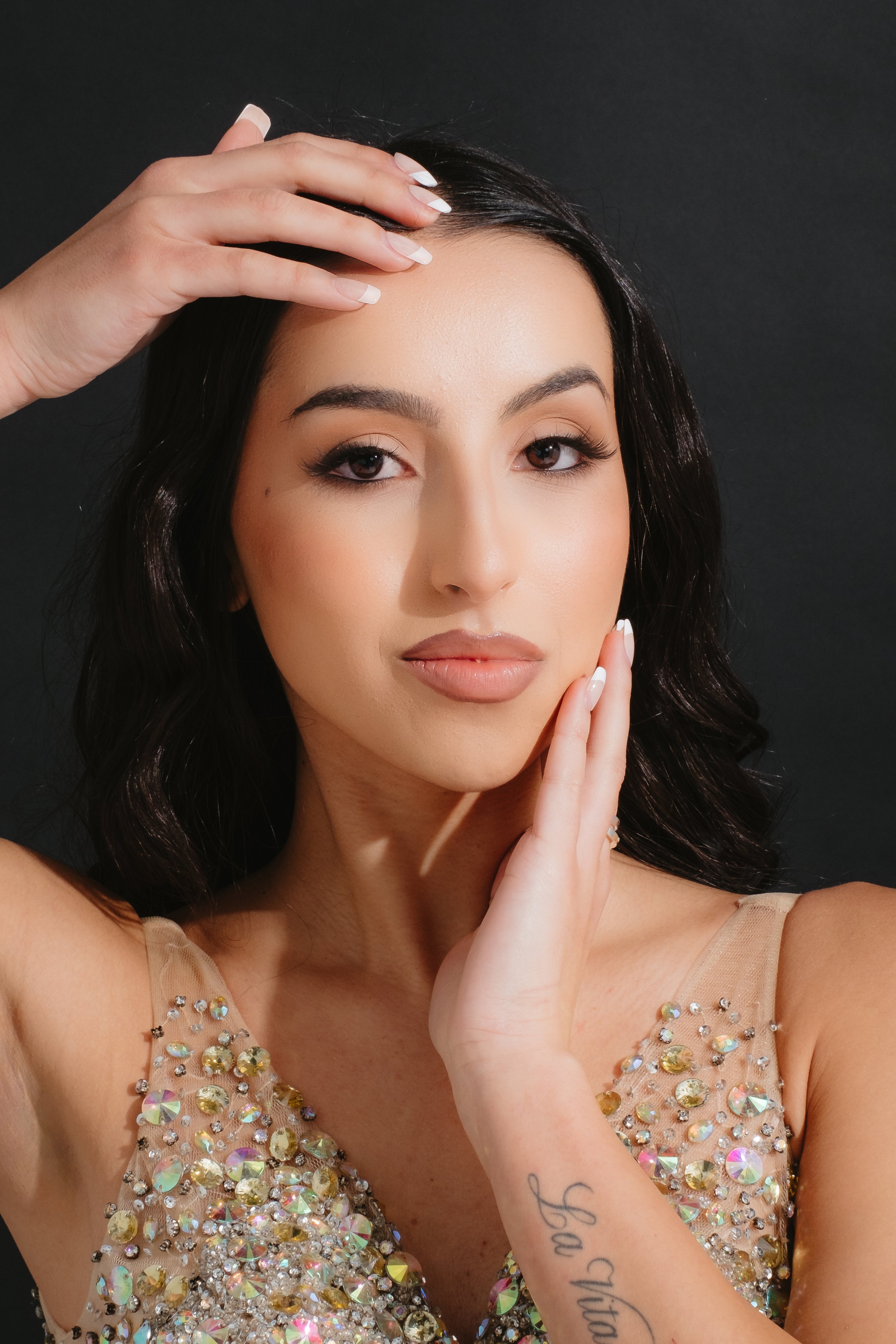 A woman with black hair and makeup wearing a sleeveless, embellished dress posing with one hand on her face and the other on her forehead against a dark background.