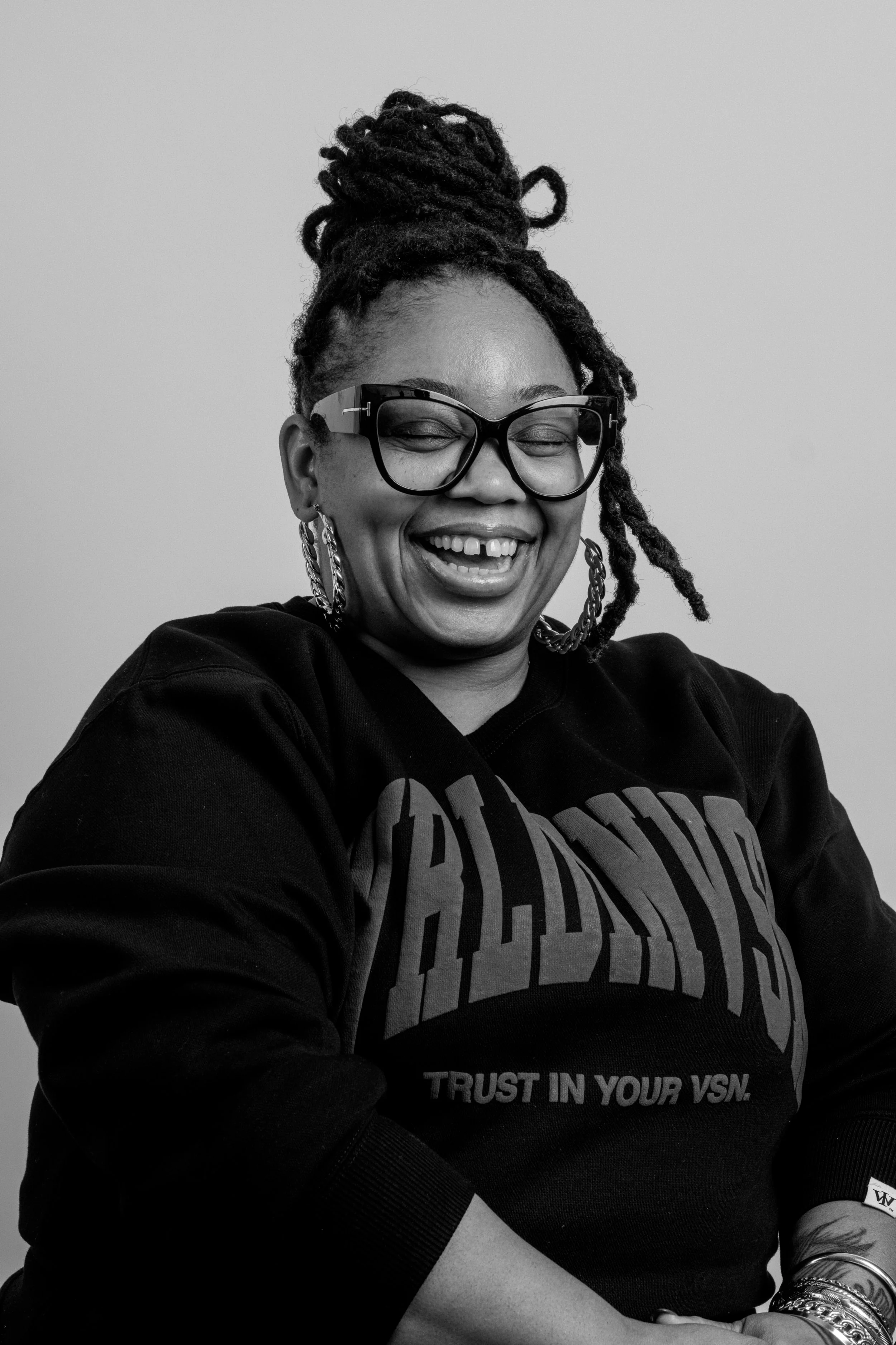 A woman with glasses and braided hair, smiling and laughing, wearing a black sweatshirt with the words "ALL DONE" and "TRUST IN YOUR VSN."