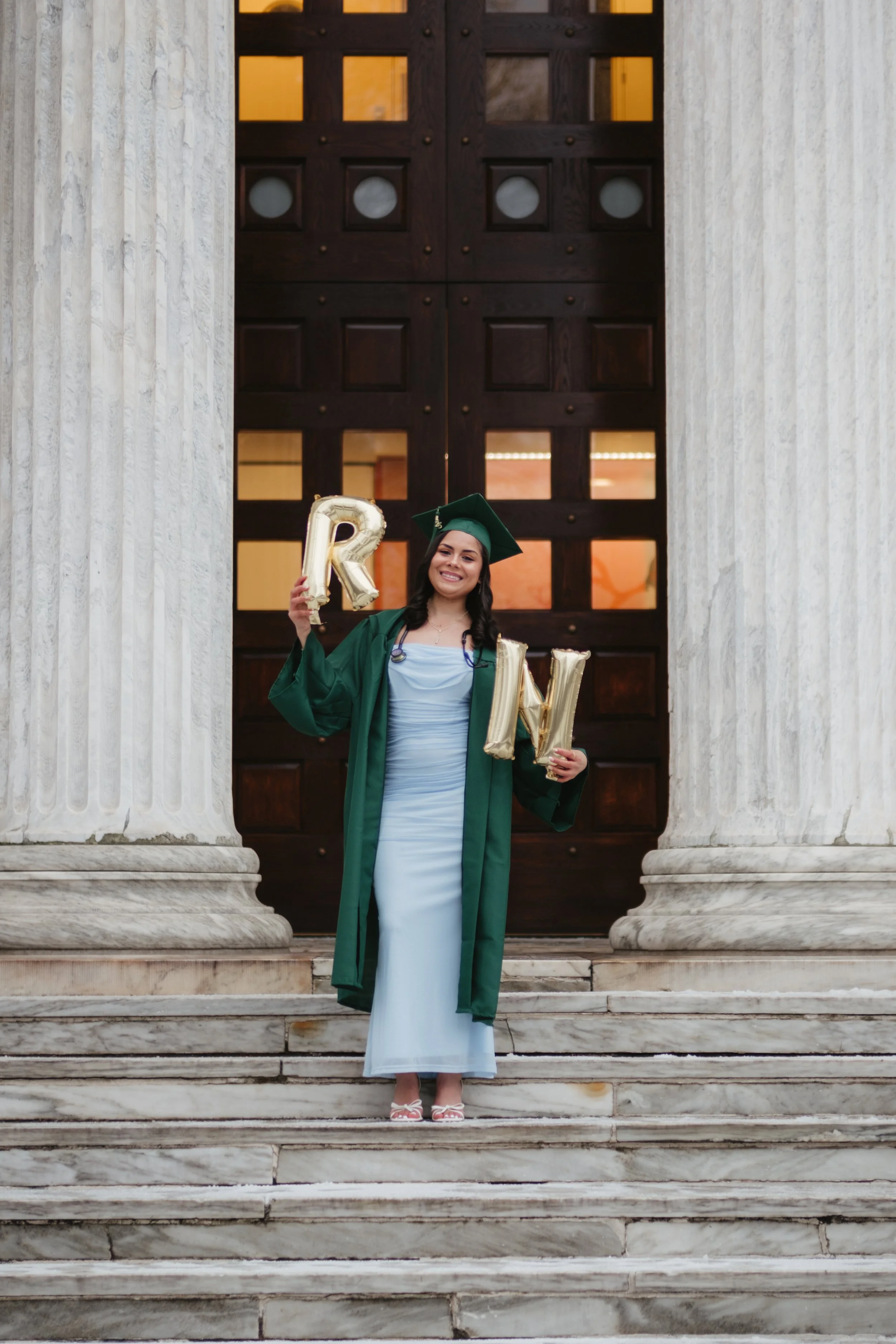 A young woman in a green graduation cap and gown standing on marble steps in front of a large wooden door, holding gold balloons spelling out 'R' and 'W', smiling at the camera.