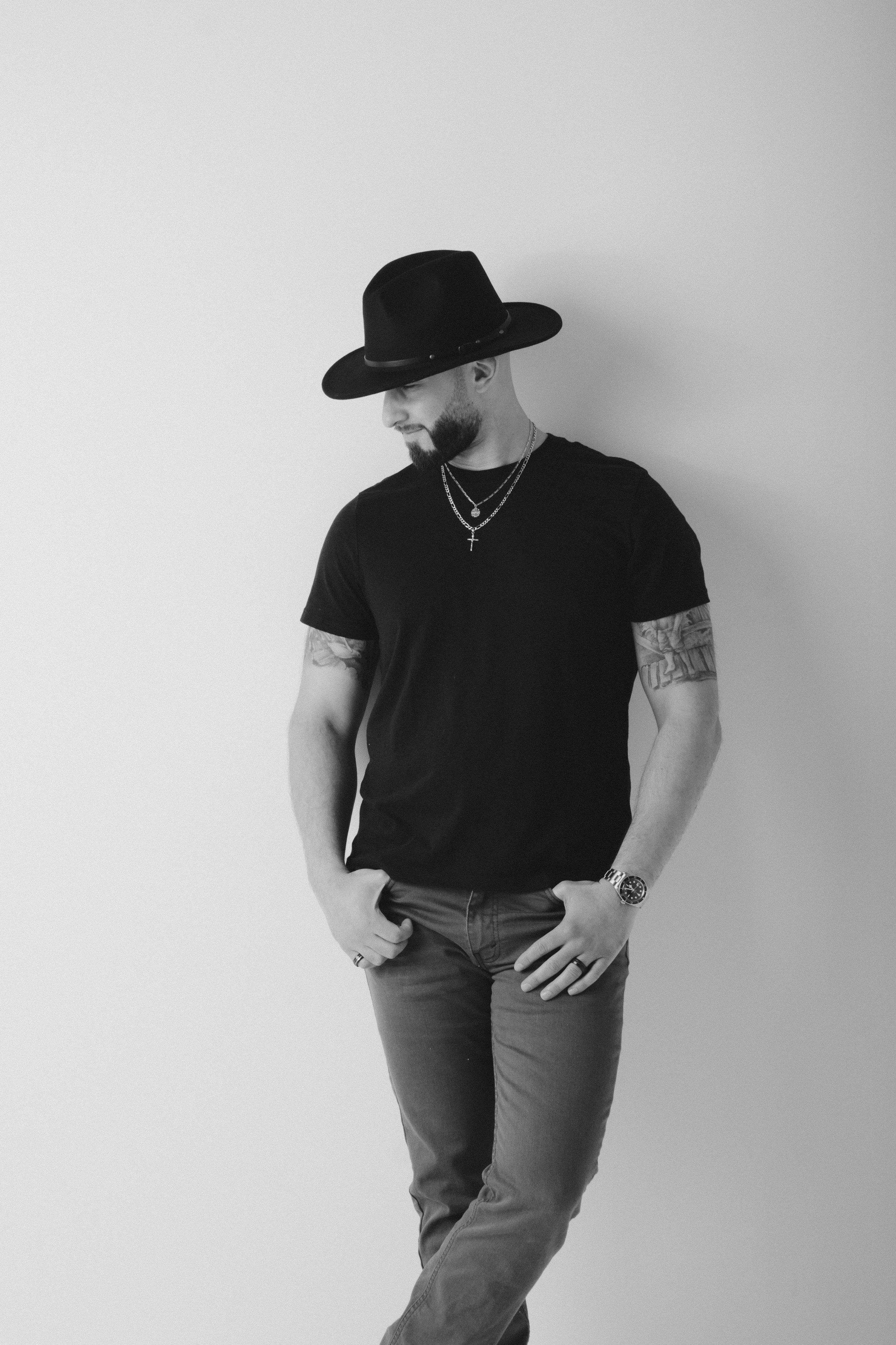 A man with a beard and tattoos, wearing a black wide-brimmed hat, black t-shirt, jeans, a watch, and layered necklaces, leaning against a plain wall with his head slightly bowed.