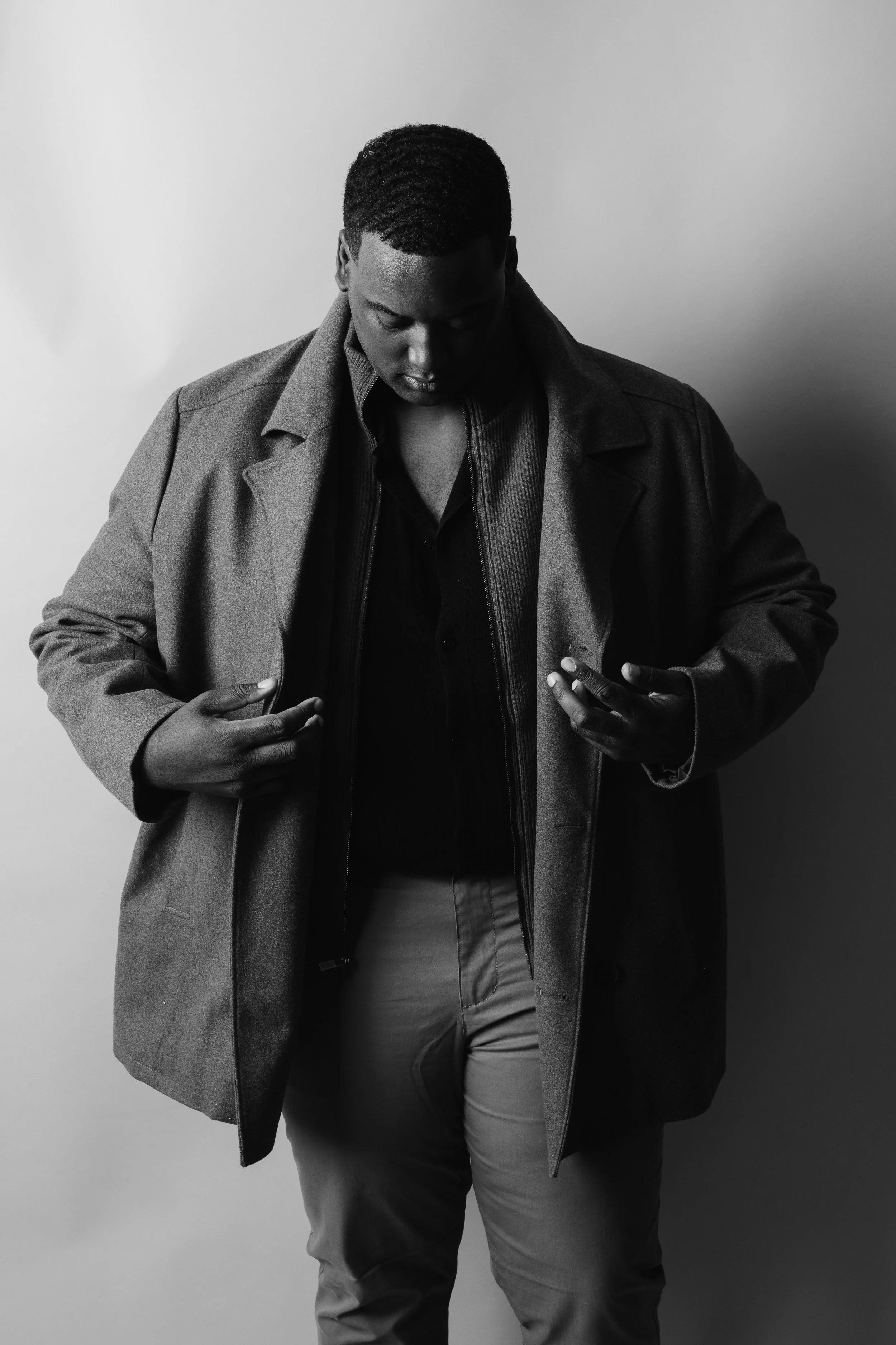 Black and white photo of a man looking at his phone, wearing a coat and casual clothes, standing against a plain background.
