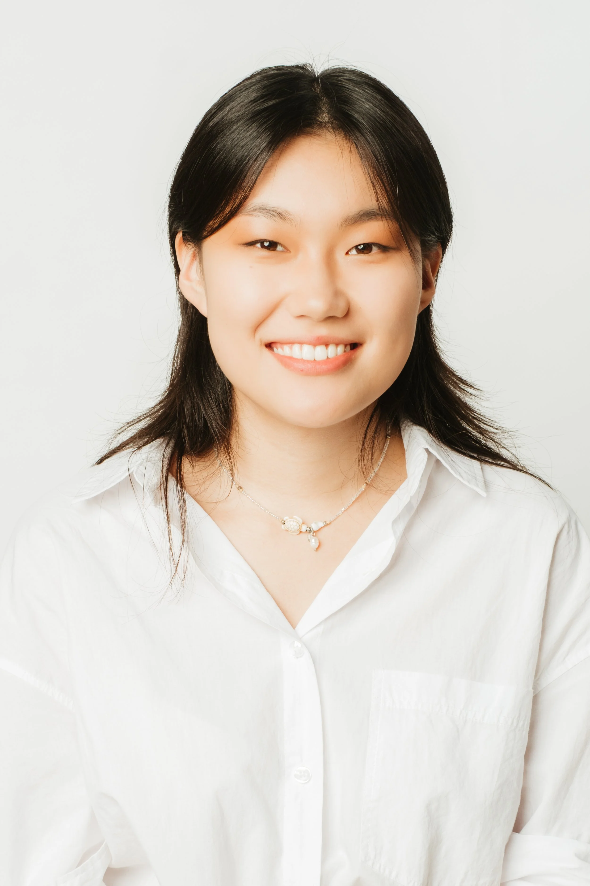 Portrait of a young woman with black hair, smiling, wearing a white shirt and a necklace, against a plain background.