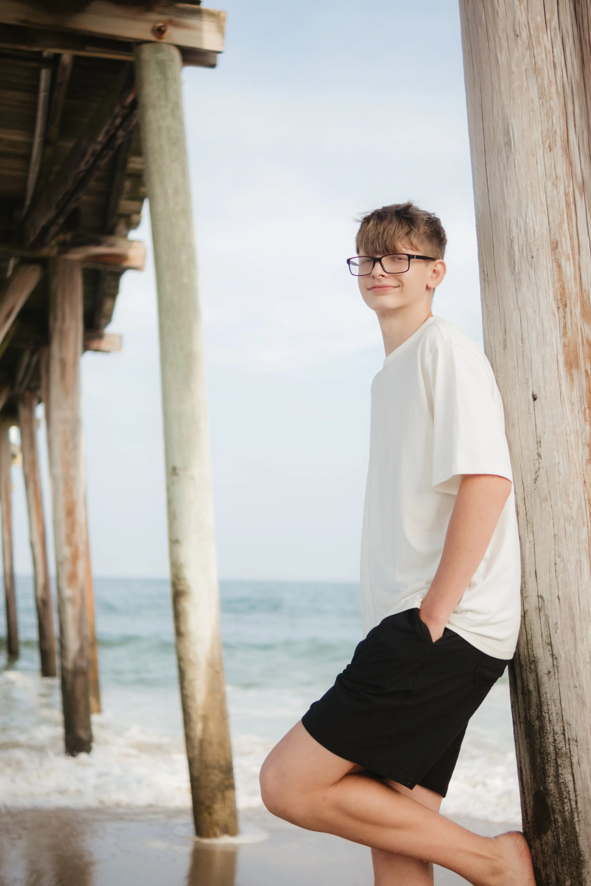 A young man with glasses, wearing a white t-shirt and black shorts, leans against a wooden pole under a pier at the beach, with the ocean and sky in the background.