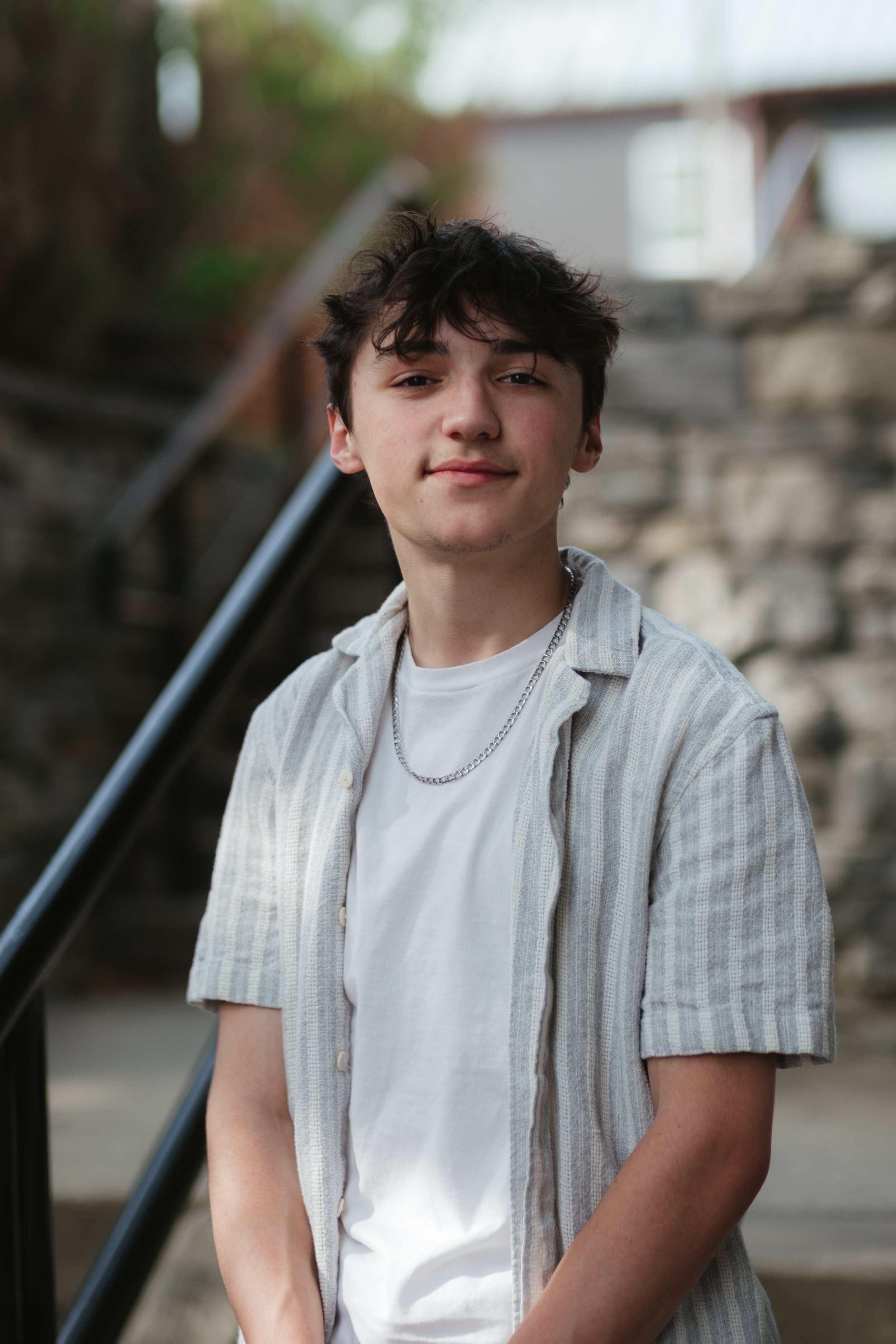A young man with dark, tousled hair and a slight smile, wearing a casual striped button-up shirt over a white t-shirt with a silver chain necklace, standing outdoors near stone steps and a metal railing.