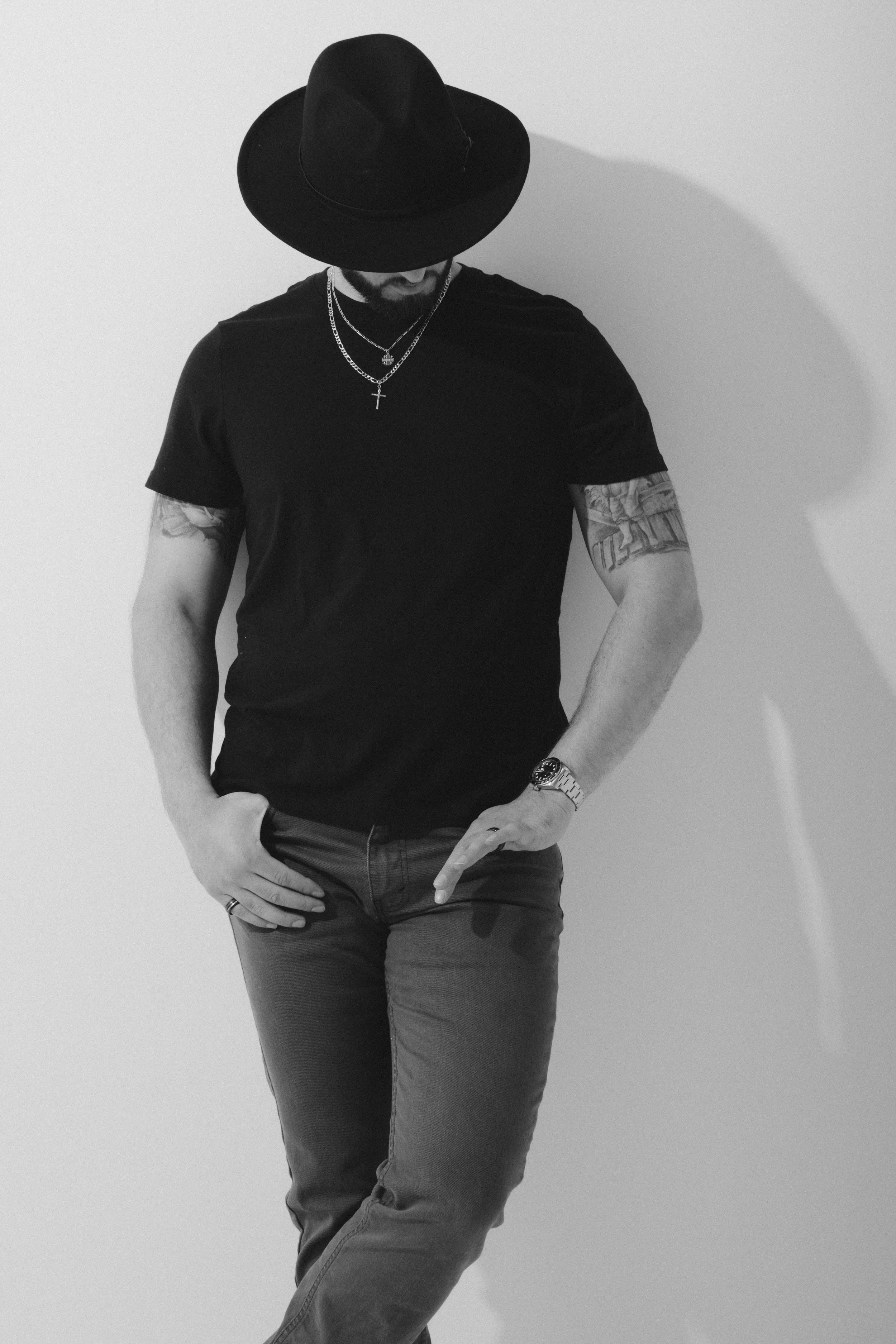 A man in a black shirt, wearing a wide-brimmed black hat, with tattoos on his arms, jewelry including a necklace, wristwatch, and ring, standing against a plain wall with a shadow cast behind him.