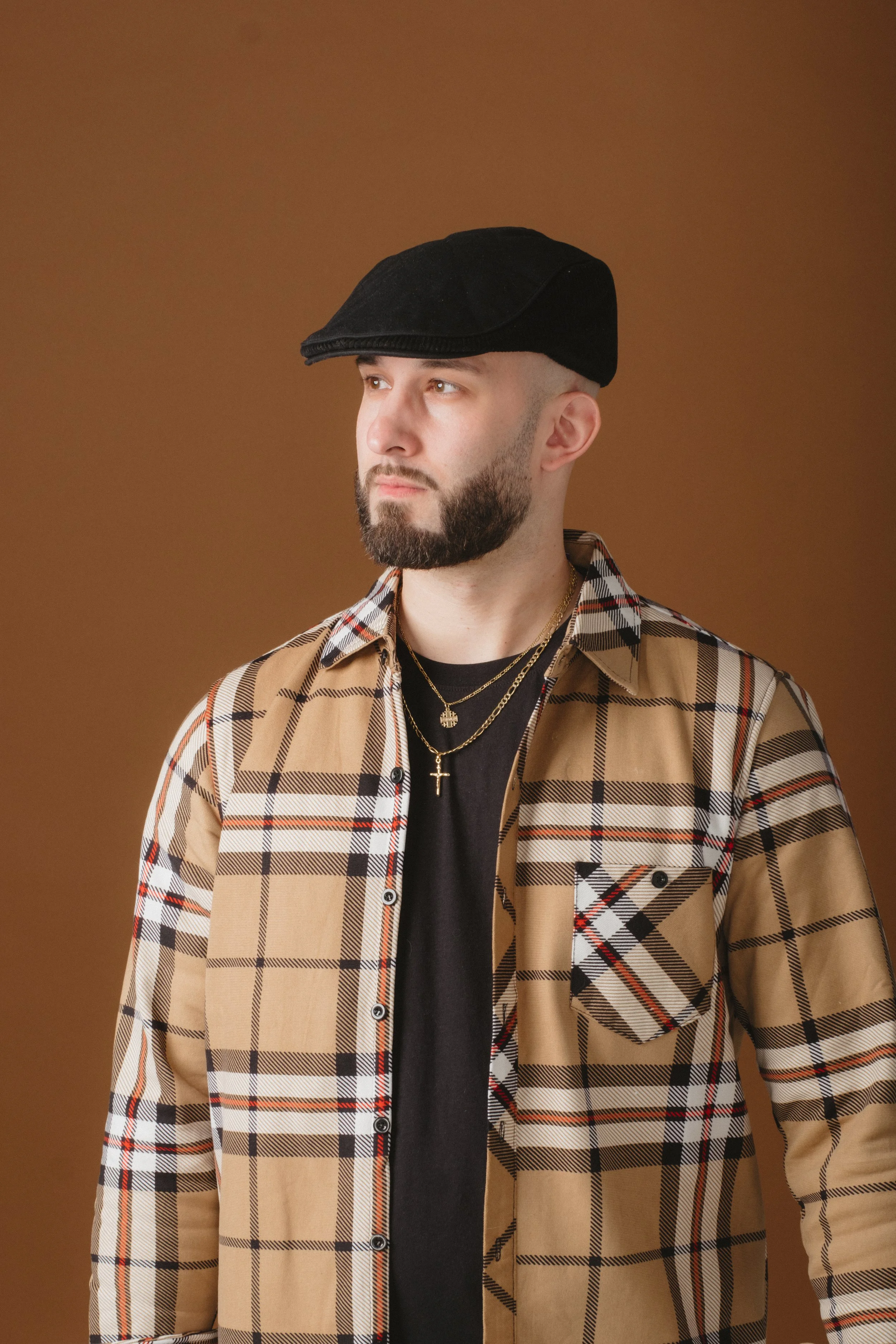 A man with a beard wearing a black flat cap, layered gold necklaces including a cross, and a beige plaid shirt over a black t-shirt, standing against a brown background.